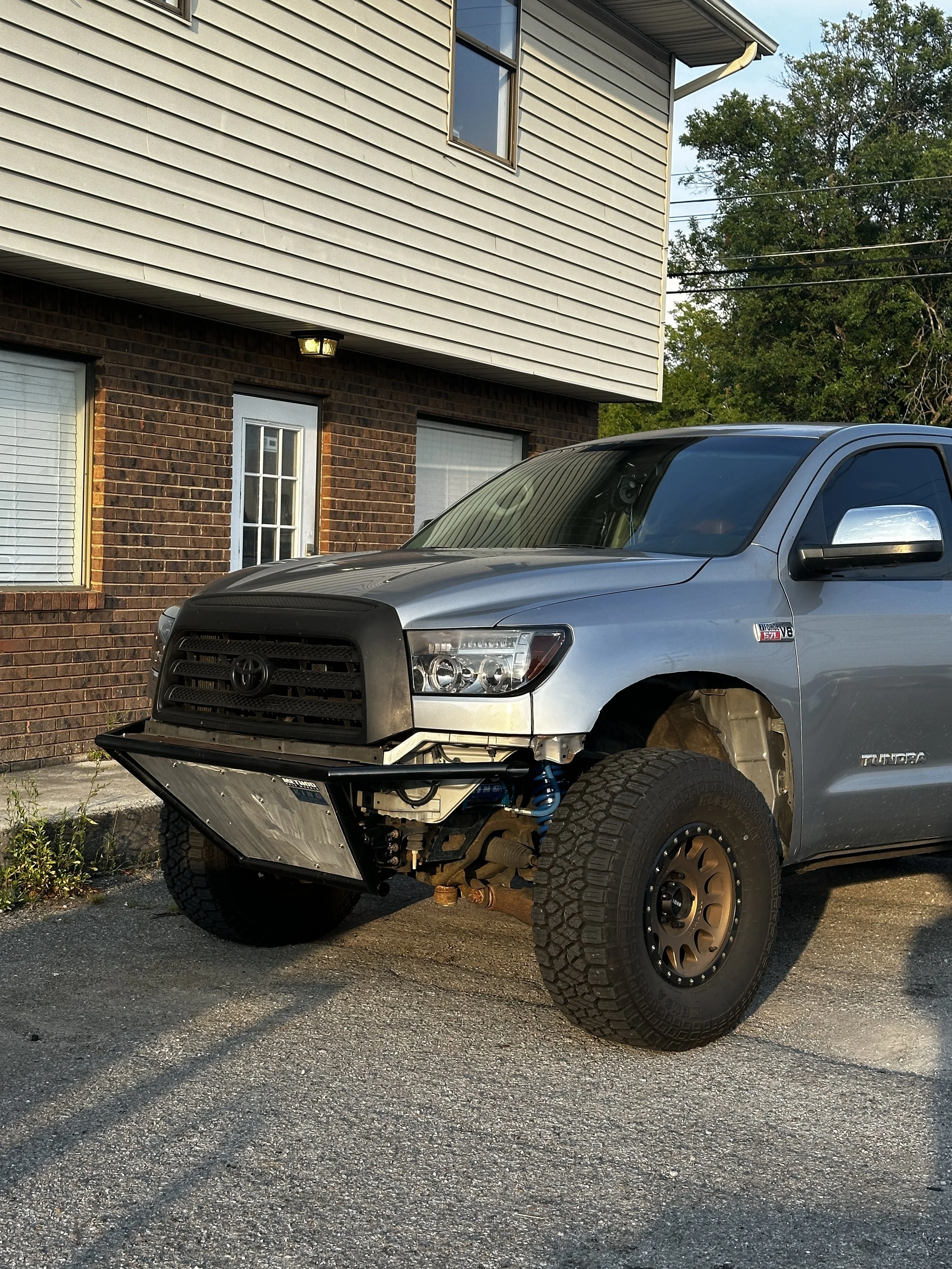 A silver Toyota Tundra pickup truck with front bumper removed, exposing the radiator and engine components, parked in front of a residential building with brick and siding exterior.