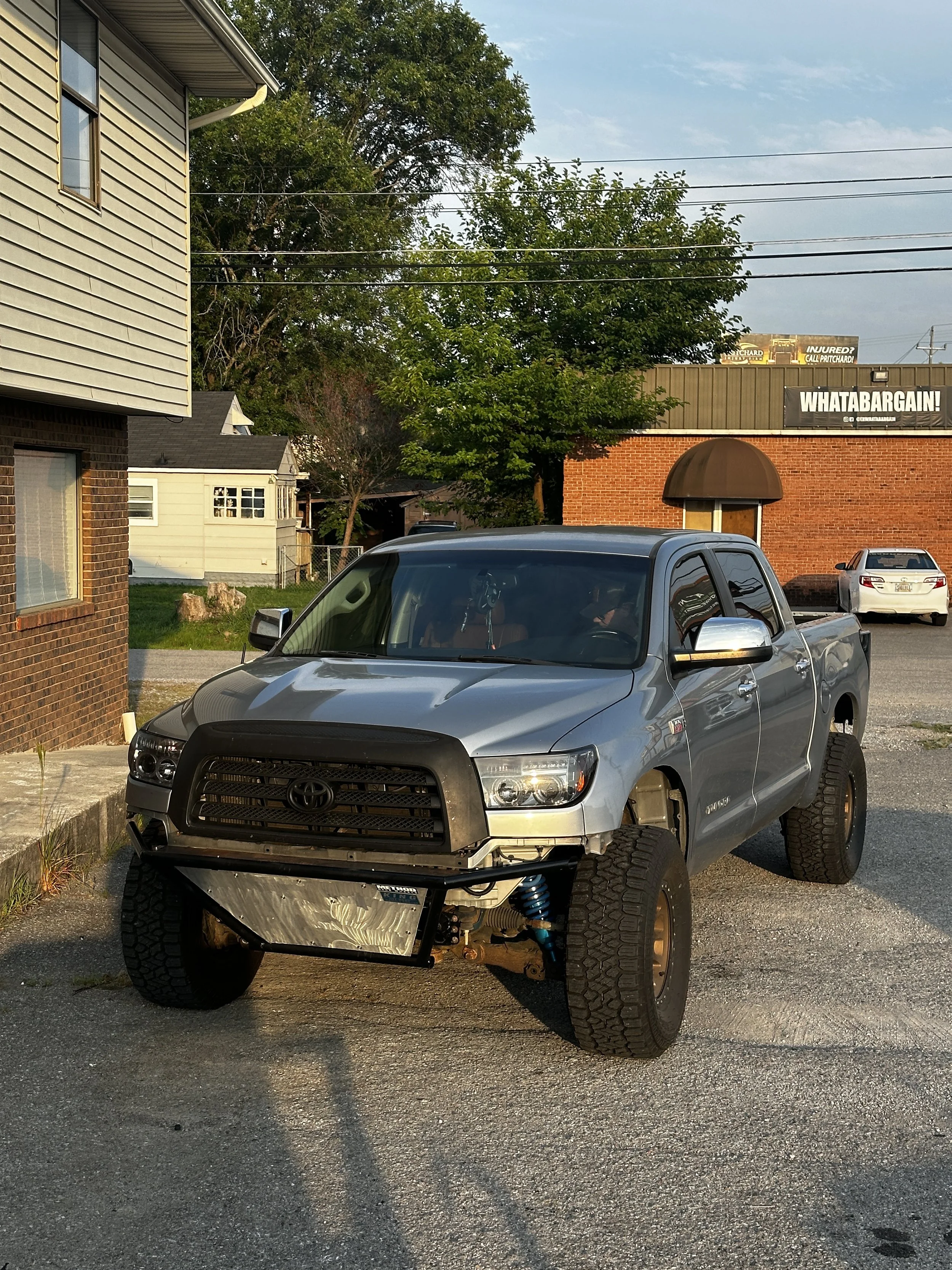 A silver Toyota pickup truck with large tires, missing front bumper, and visible suspension components parked on a gravel lot near a brick building, with trees and other cars in the background.