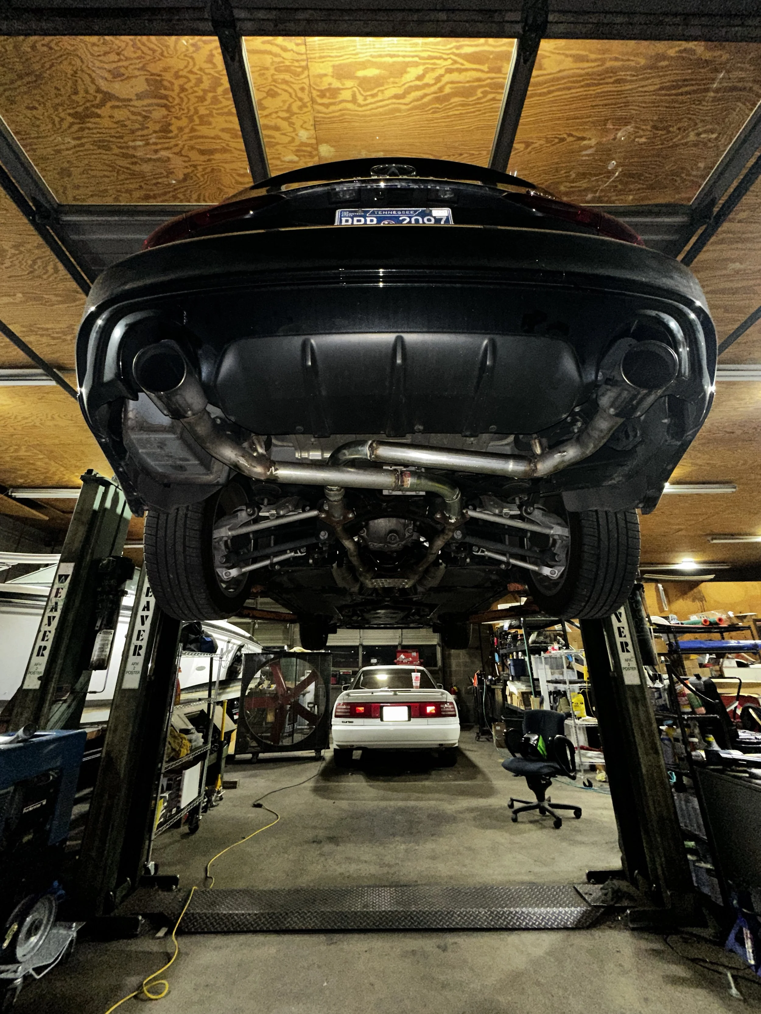 The image shows the underside of a black car lifted on a hydraulic lift in a garage. There is another white car parked inside the garage, along with various tools, equipment, and shelves.