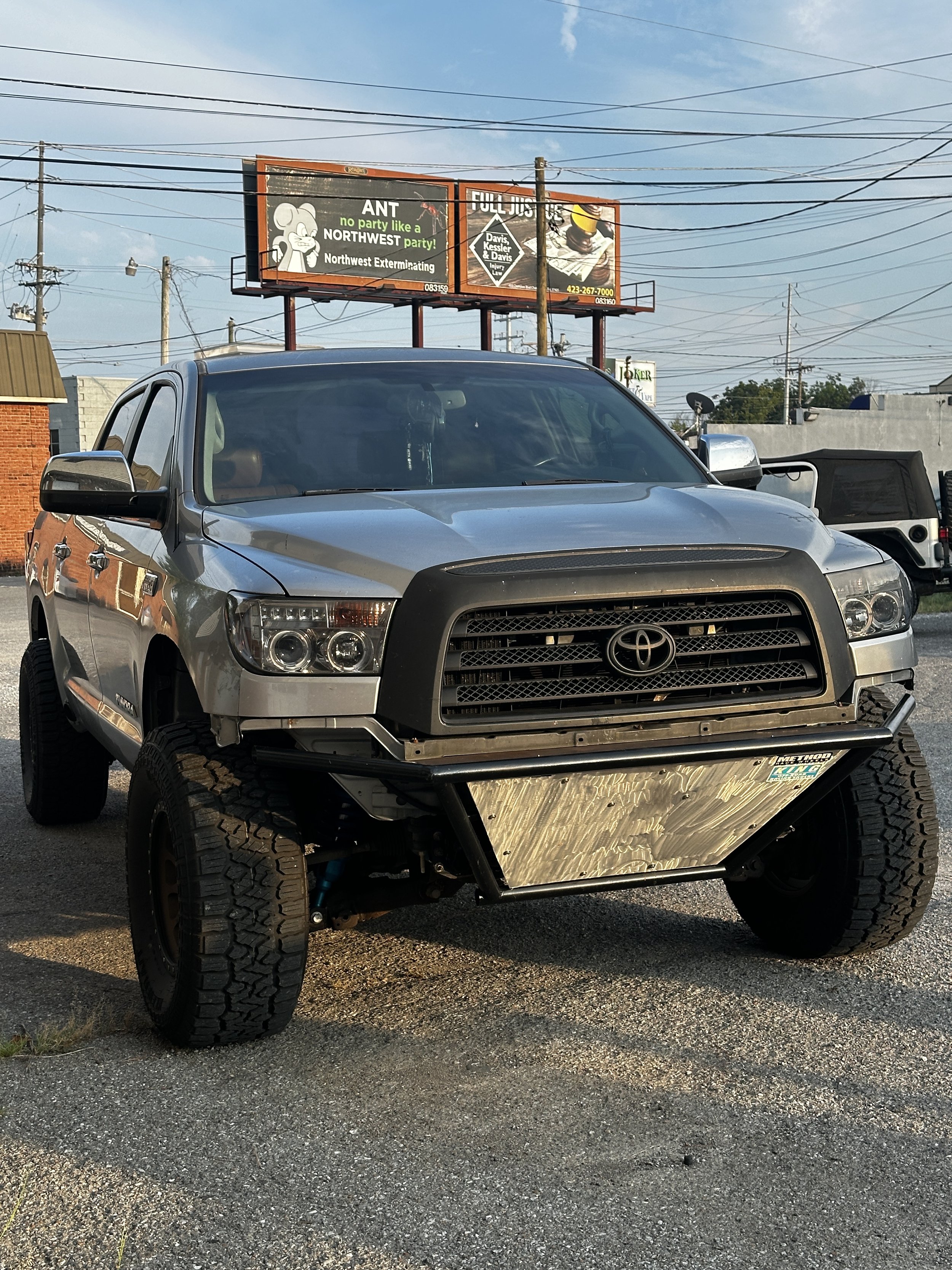 A silver Toyota truck with a modified front bumper and off-road tires parked outdoors in a lot, with overhead power lines and a billboard sign in the background.