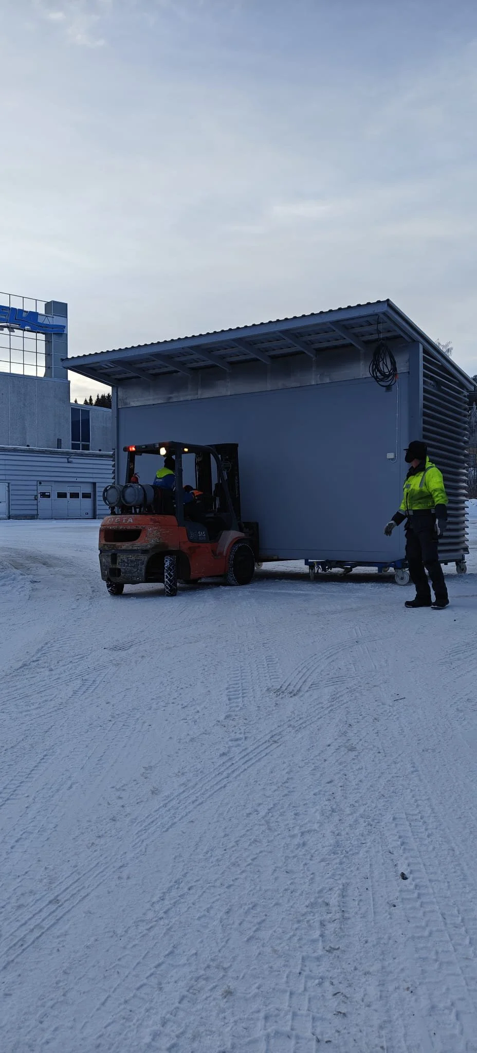 Kuvassa on kaksi työntekijää siirtämässä suurta rakennusosaa, ympärillä on luminen maa.