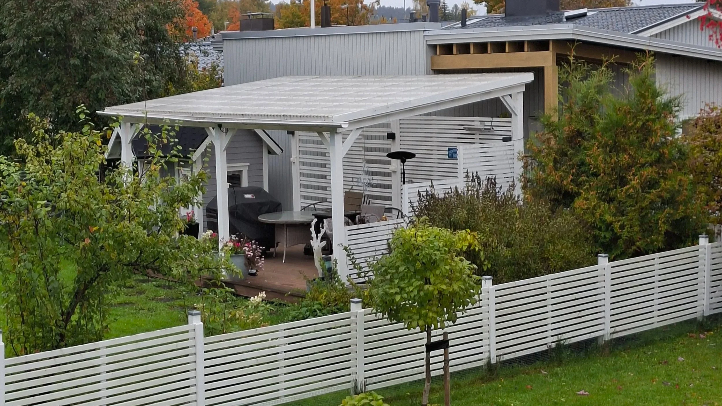 Kattoterassi takapihalla, jossa pöytä ja tuoleja, grillilaitteita, ympäröivät puita ja pensaita, ja valkoinen aita.