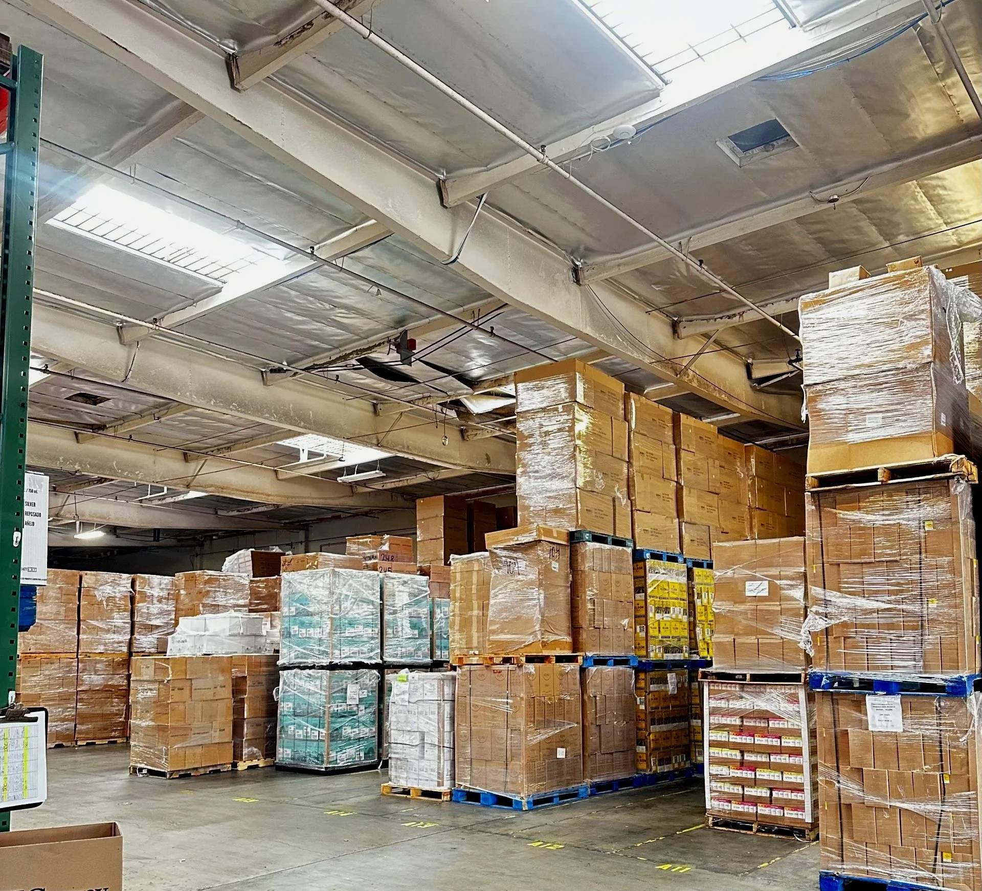 Warehouse with stacked pallets of boxed goods wrapped in plastic, organized on metal shelves and on the floor.
