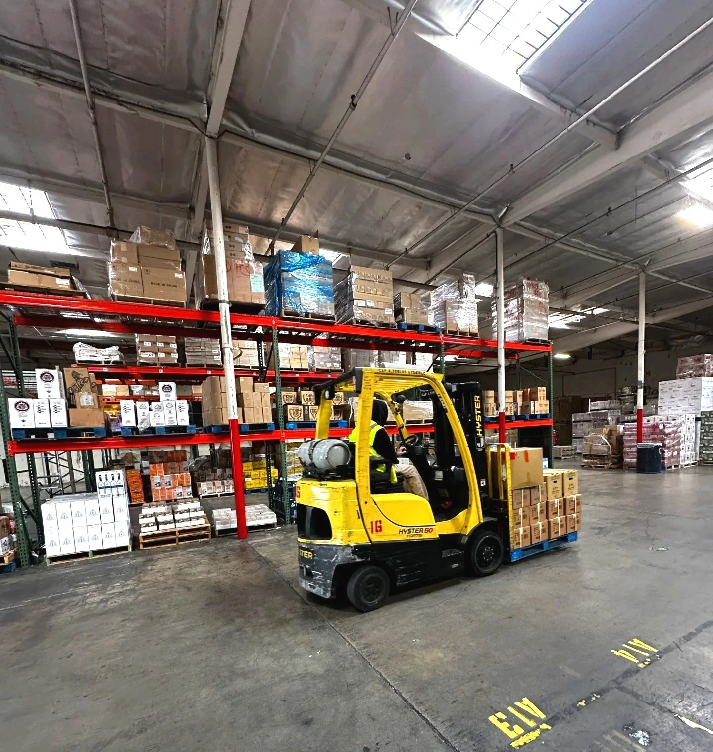 A warehouse with a yellow forklift moving pallets of boxes and products, with shelves stacked with various boxed goods along the wall.