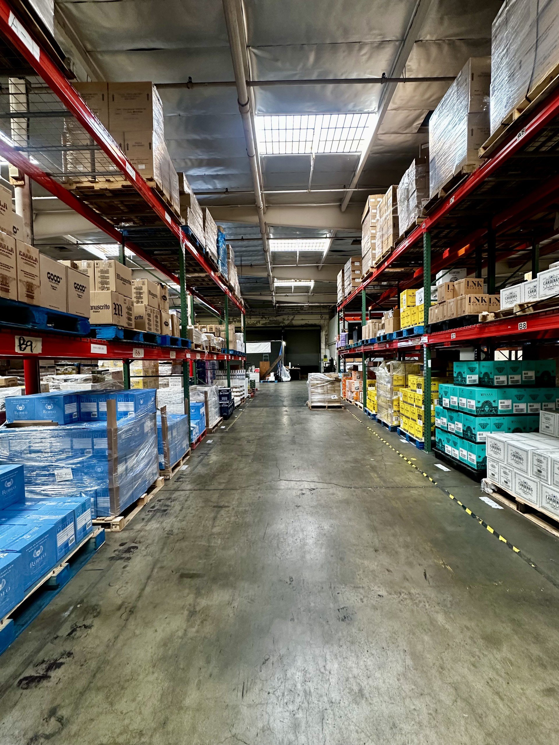 A view down the aisle of a warehouse with industrial shelves stacked with boxes and pallets.