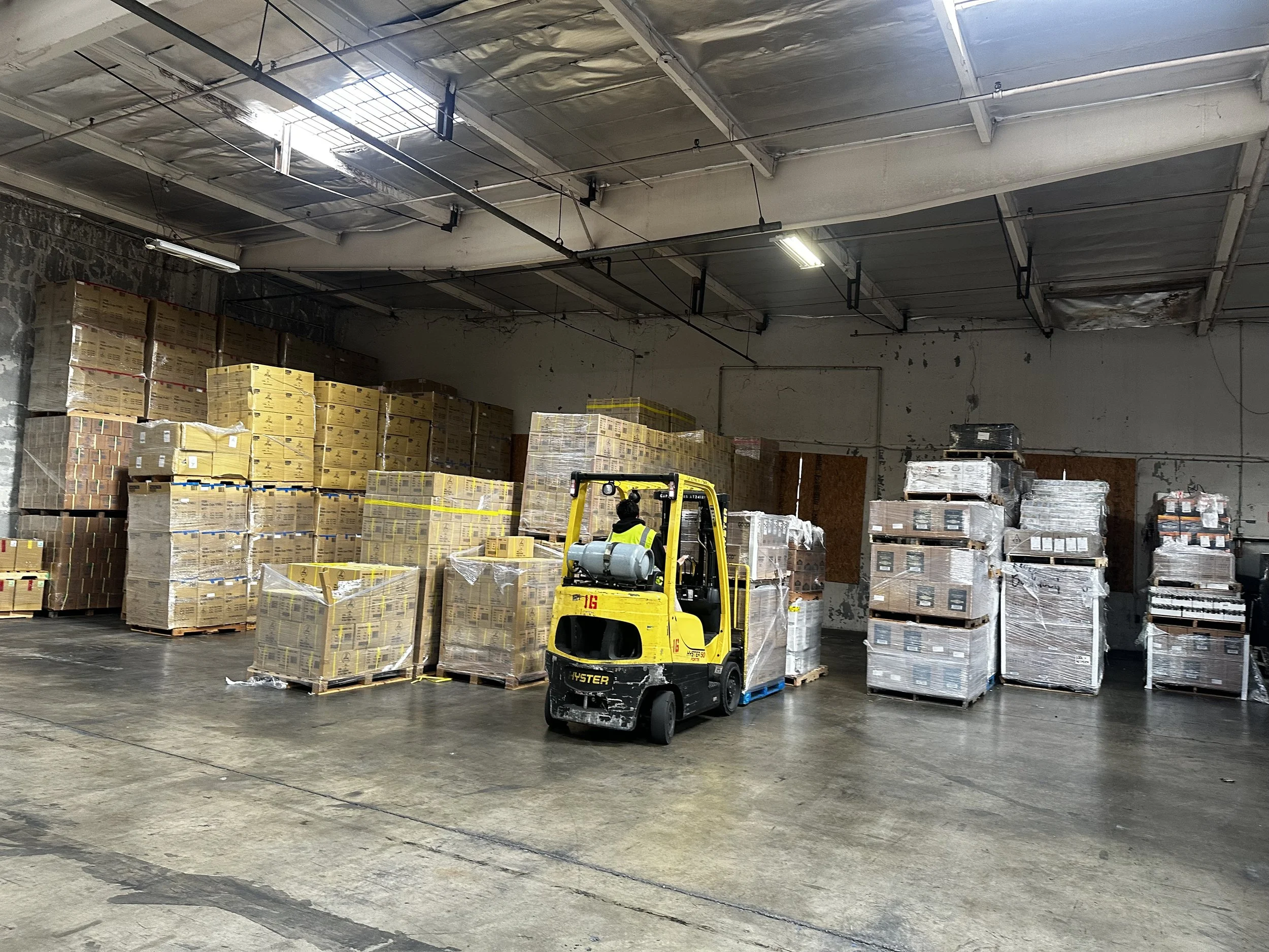 Inside a warehouse with a forklift moving pallets of boxed goods. The warehouse has a concrete floor, high ceiling with skylights, and walls showing signs of wear.