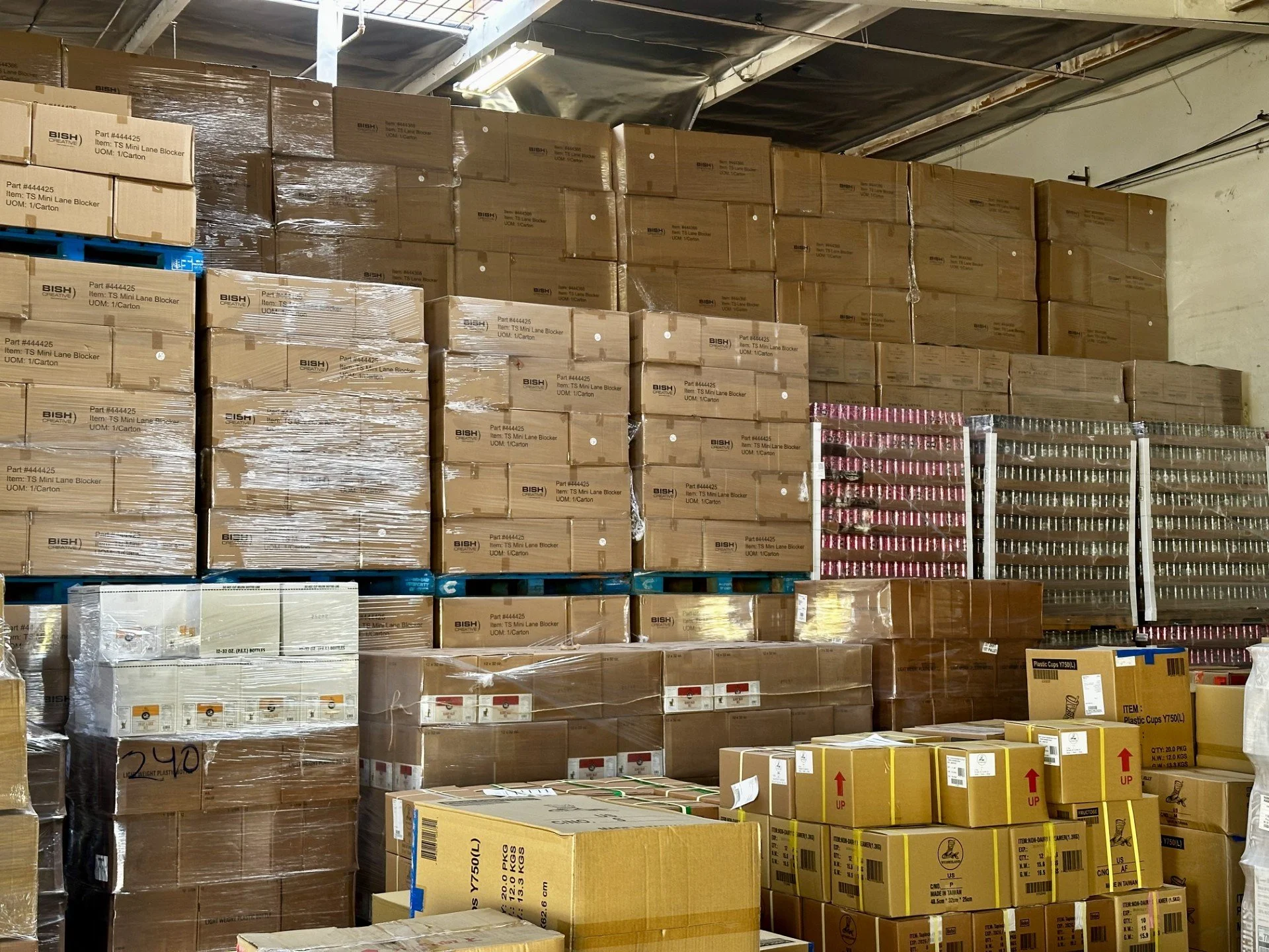 A storage warehouse with stacked cardboard boxes and plastic containers on shelves and the floor, some wrapped in plastic, in a warehouse setting with ceiling lights.