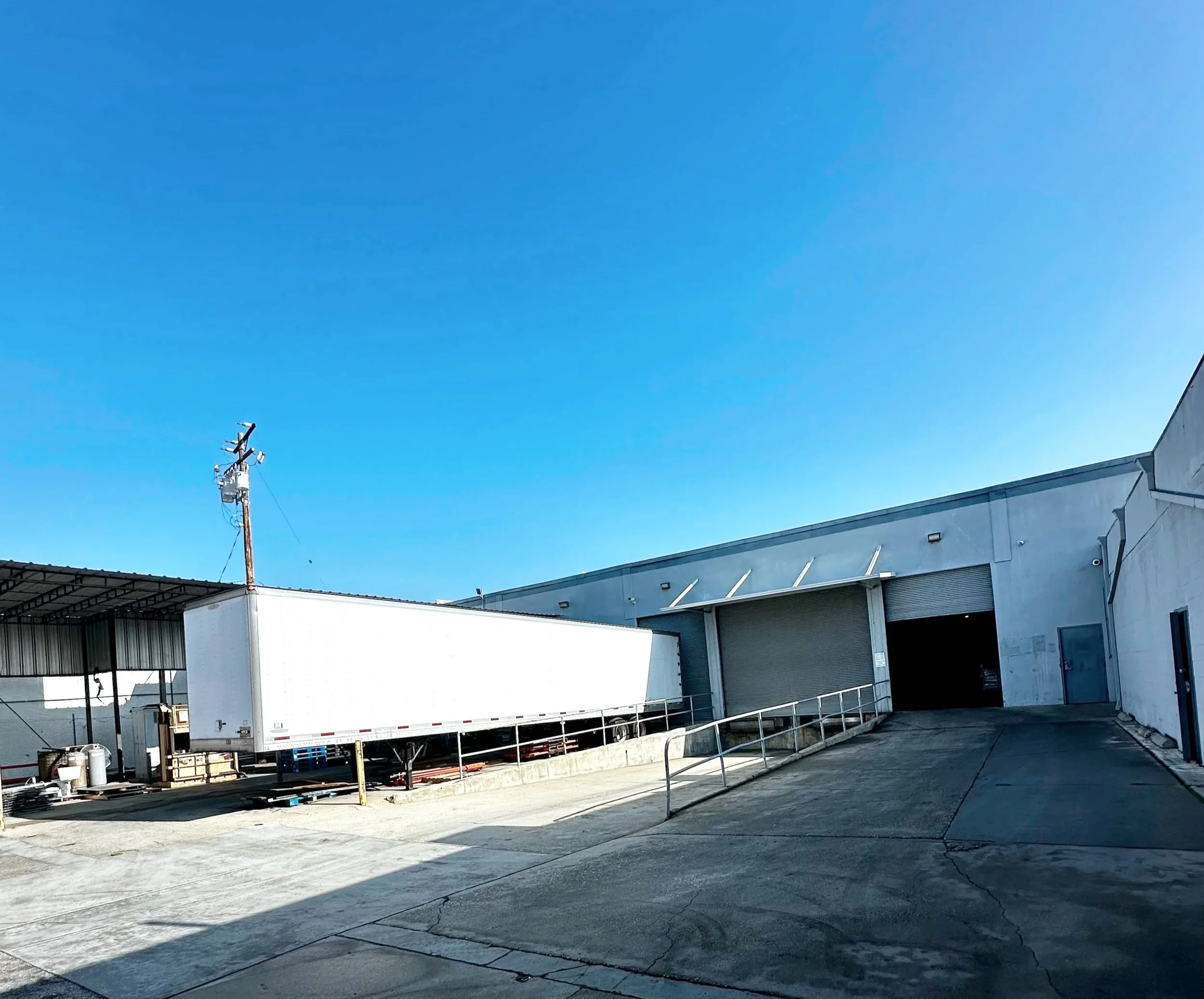 Loading dock with a large white truck trailer and warehouse buildings under a clear blue sky.