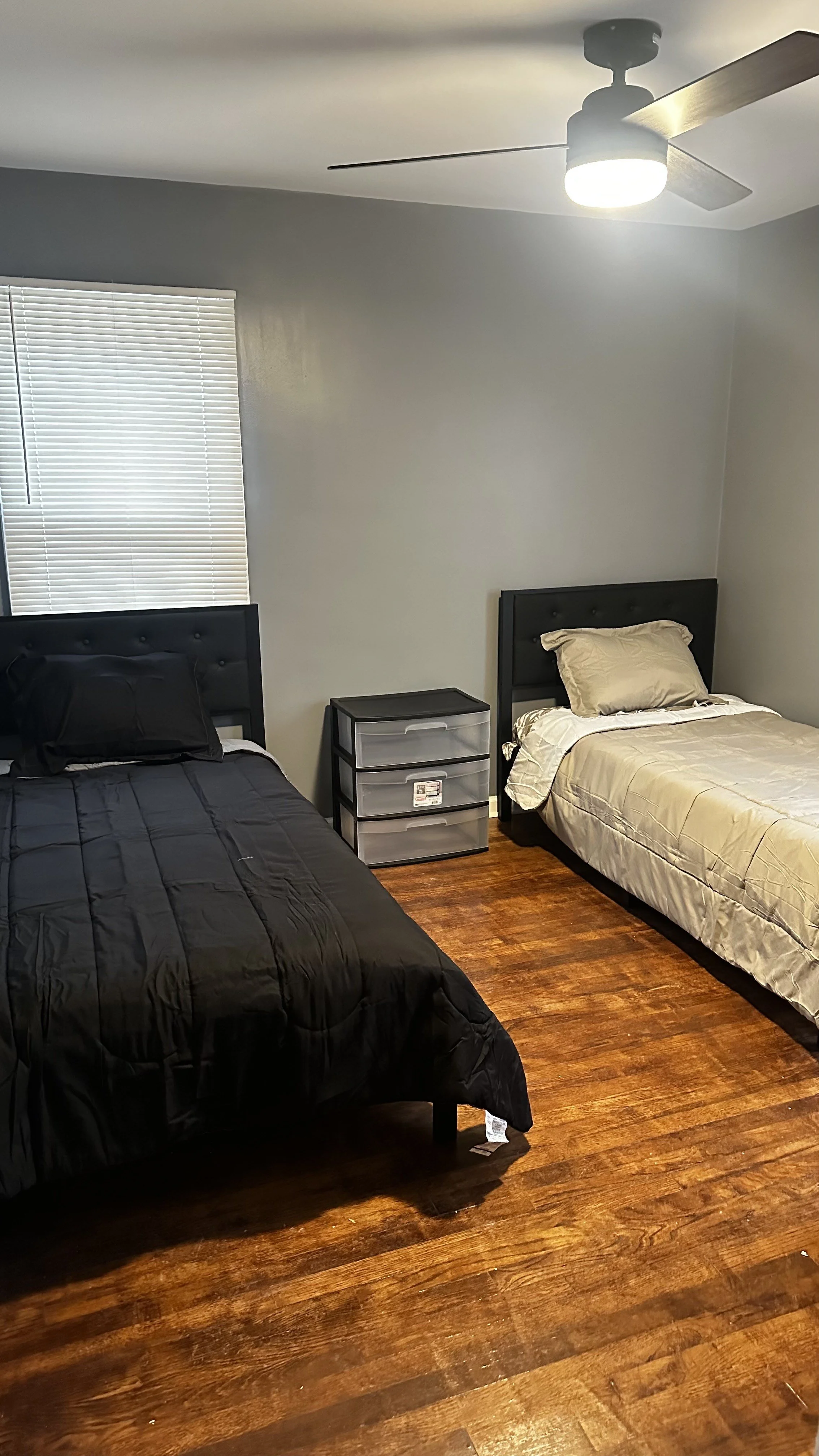 Two single beds with black and beige bedding, a plastic drawer storage bin between them, a window with closed blinds, hardwood flooring, and a ceiling fan with light.