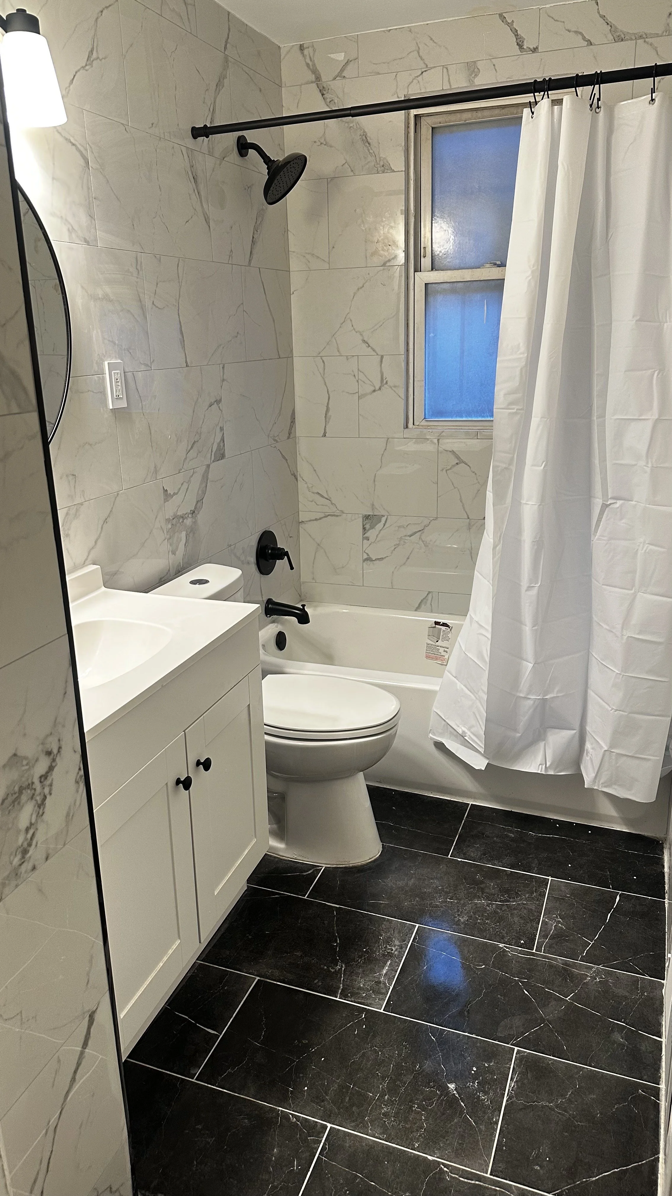 A modern bathroom with marble tiled walls, a bathtub with a black showerhead, a small sink with a white cabinet, a toilet, a small window, and a white shower curtain.