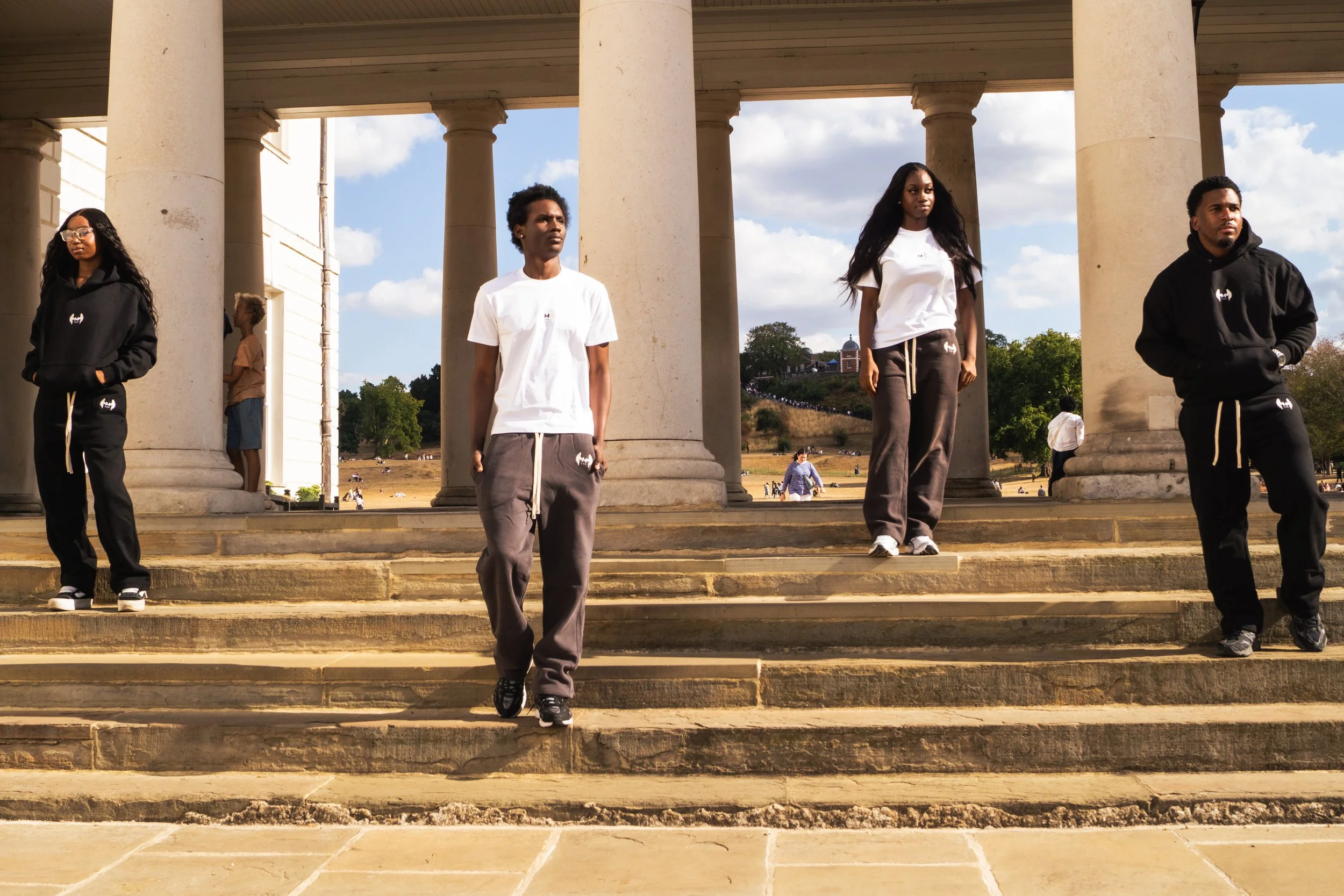 Four young people standing on steps in front of a large columned building, outdoors on a sunny day, with a park and blue sky in the background. Two are wearing black hoodies and sweatpants, and two are wearing white t-shirts with grey sweatpants.