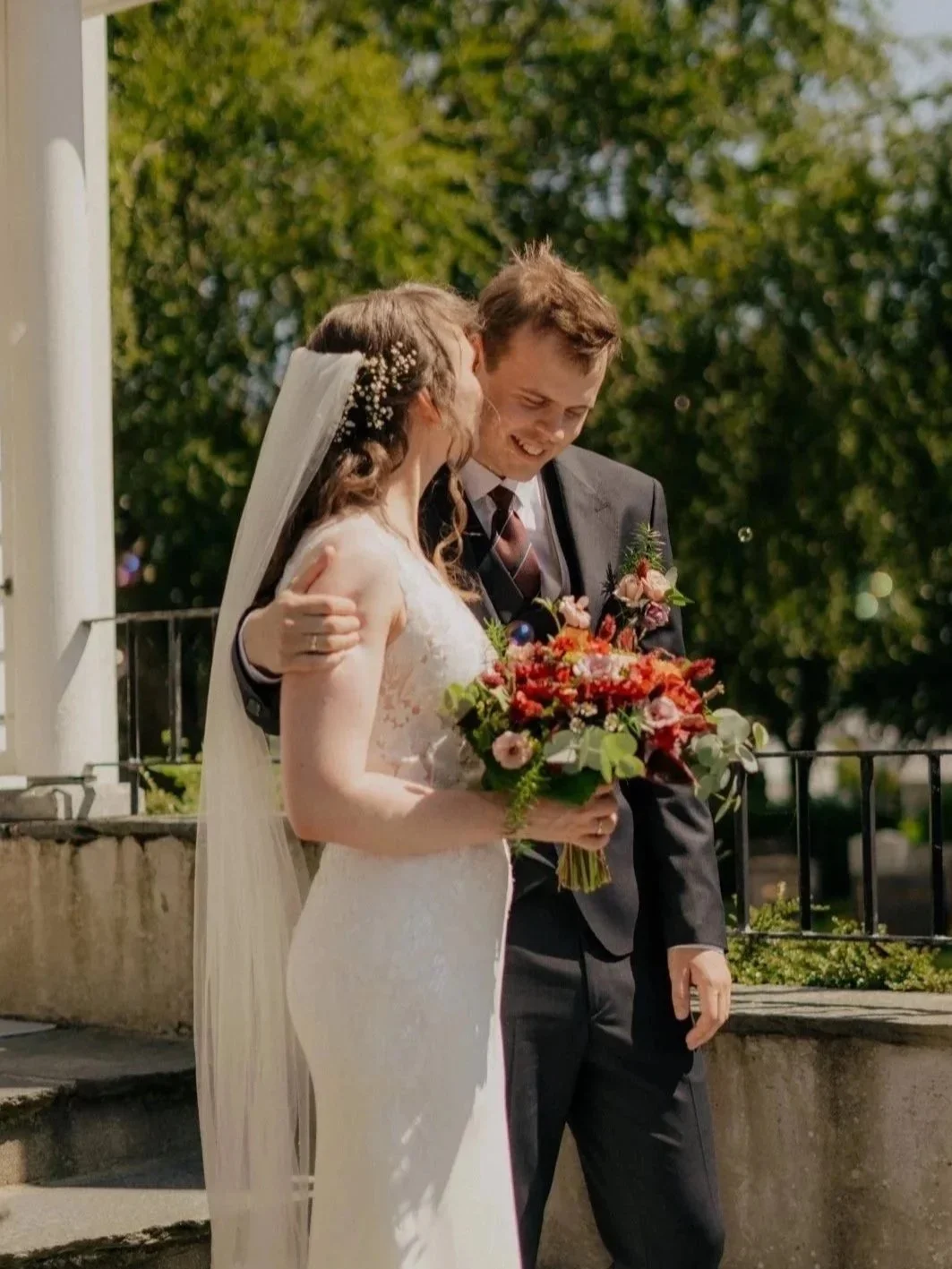 Et brudepar som kysser, kvinnen holder en bukett med blomster, ute i solen med grønne trær i bakgrunnen.