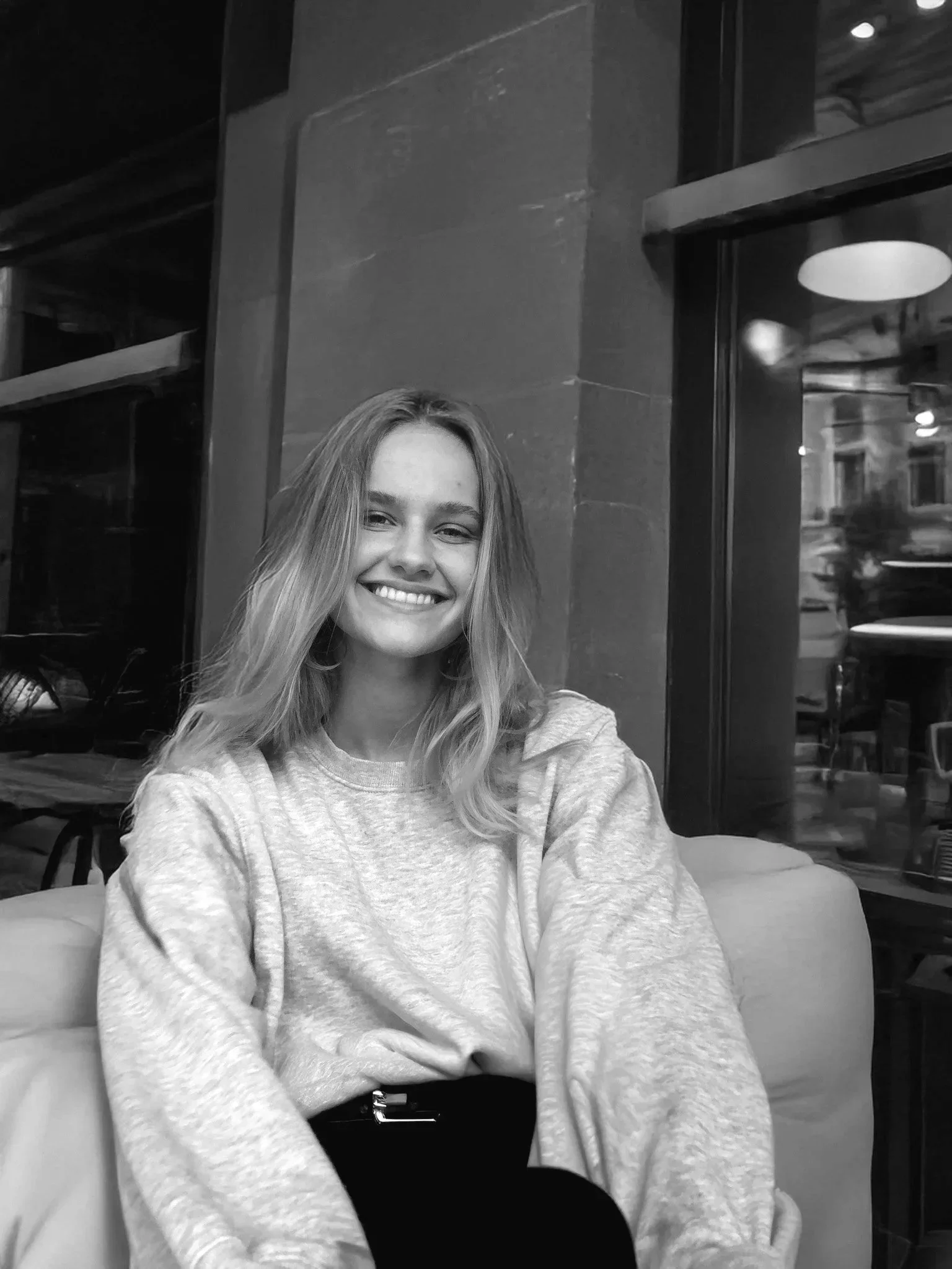 Et bilde av en smilende jente som sitter i en kafé, kledd i en lys genser og svarte bukser, foran store vinduer.