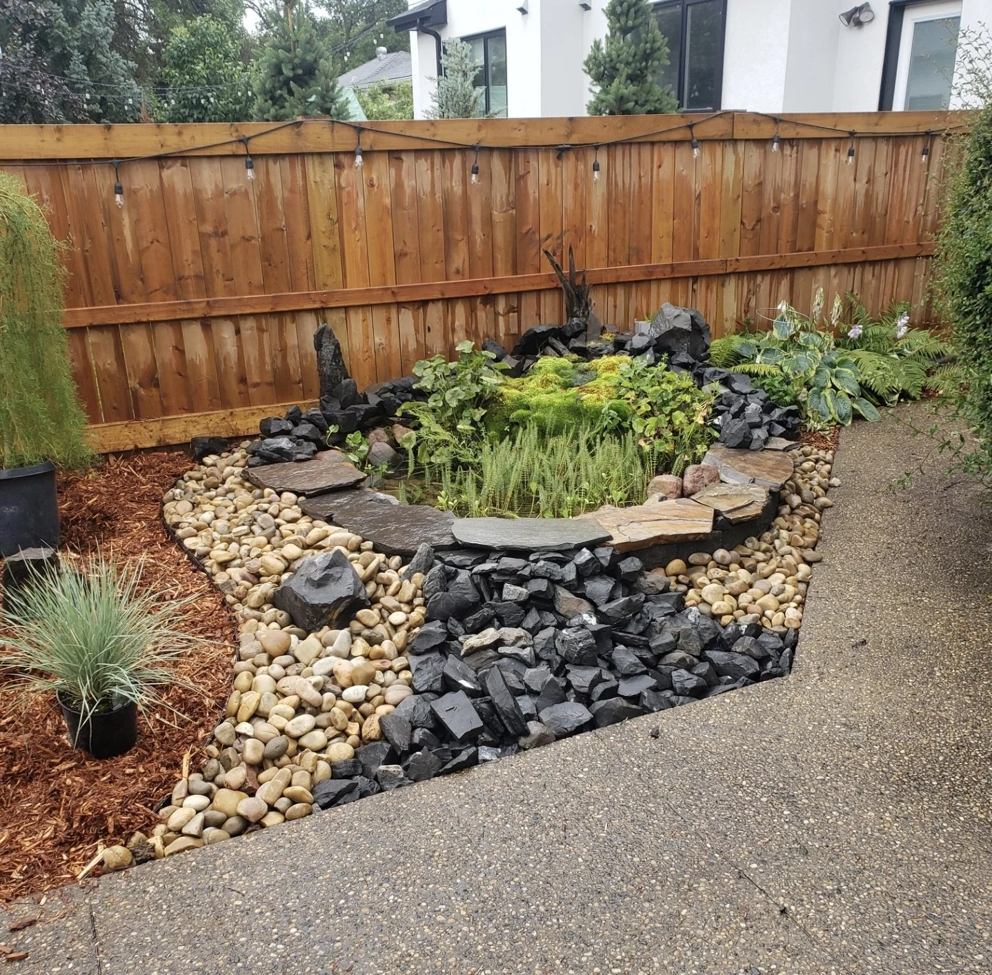 Decorative rock garden bed with river rock, black slate, and flagstone caps beside a wooden fence with string lights