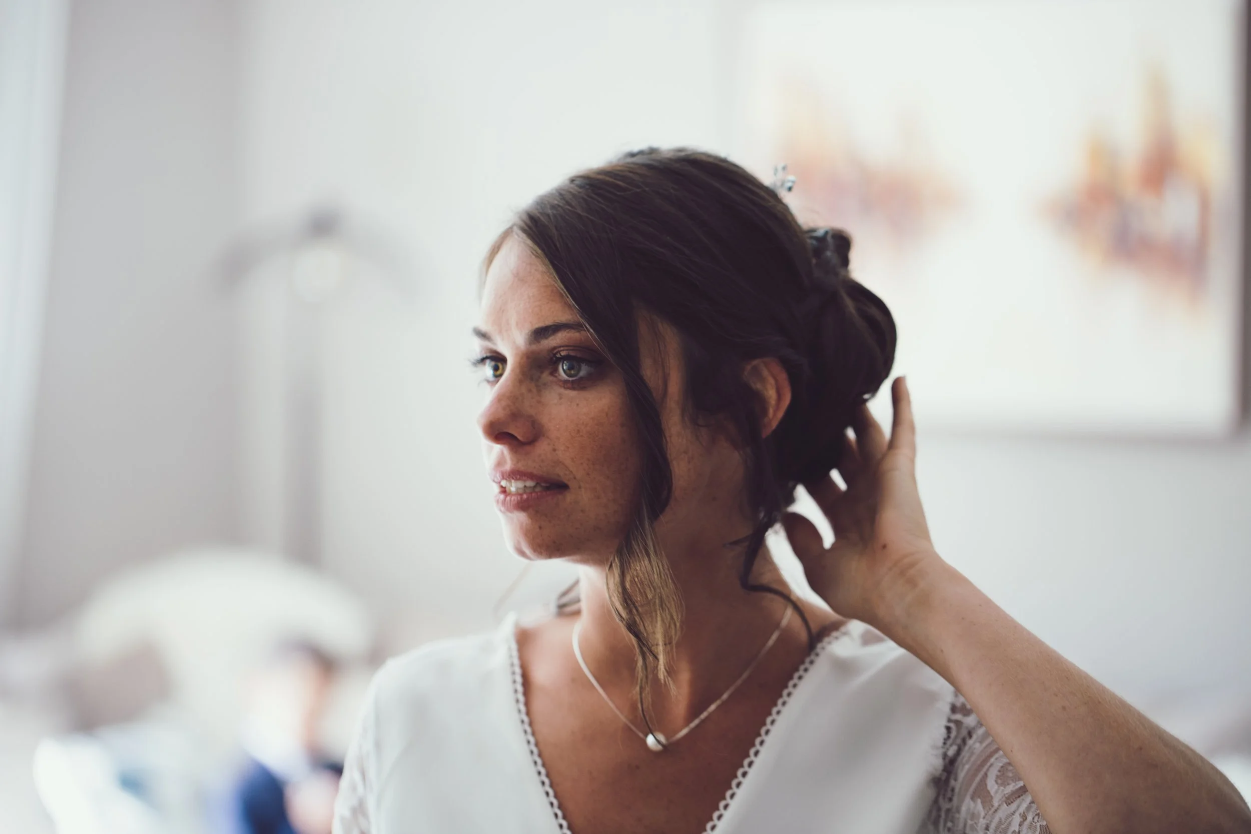 Une femme avec des cheveux bruns et des yeux bleus ajuste sa coiffure dans un miroir, portant une robe blanche