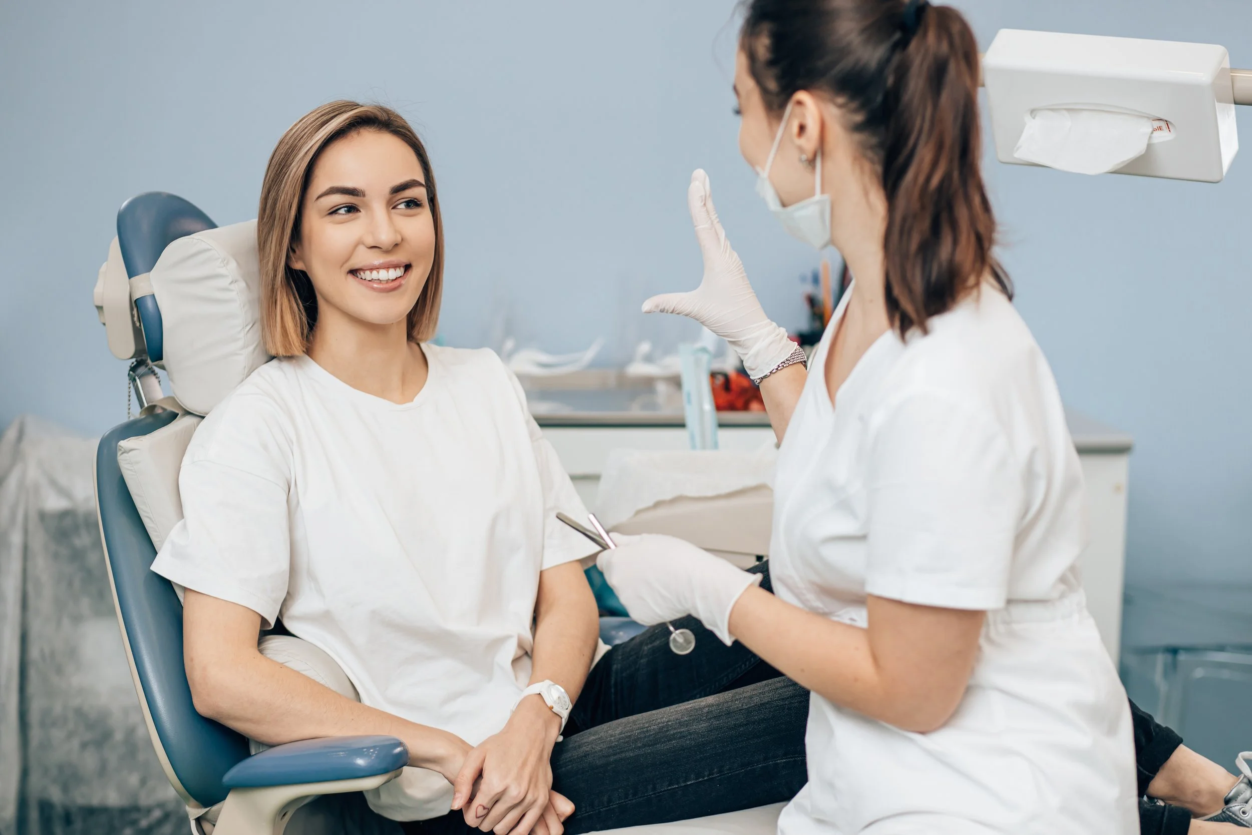 A young woman sitting in a dental chair smiling at her dentist, who is wearing a mask and gloves, and appears to be explaining or discussing dental care.