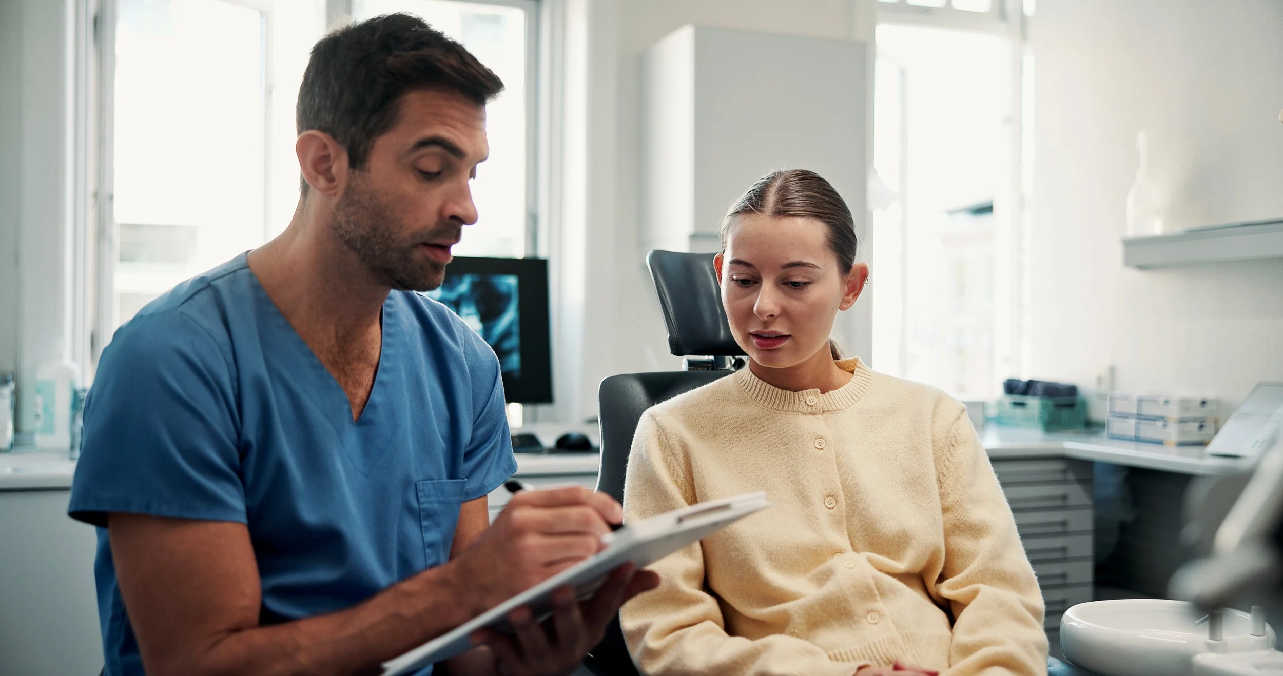 A male doctor or nurse in blue scrubs sitting next to a young woman in a medical office or clinic. The doctor is writing on a notepad, and the woman looks at him attentively.