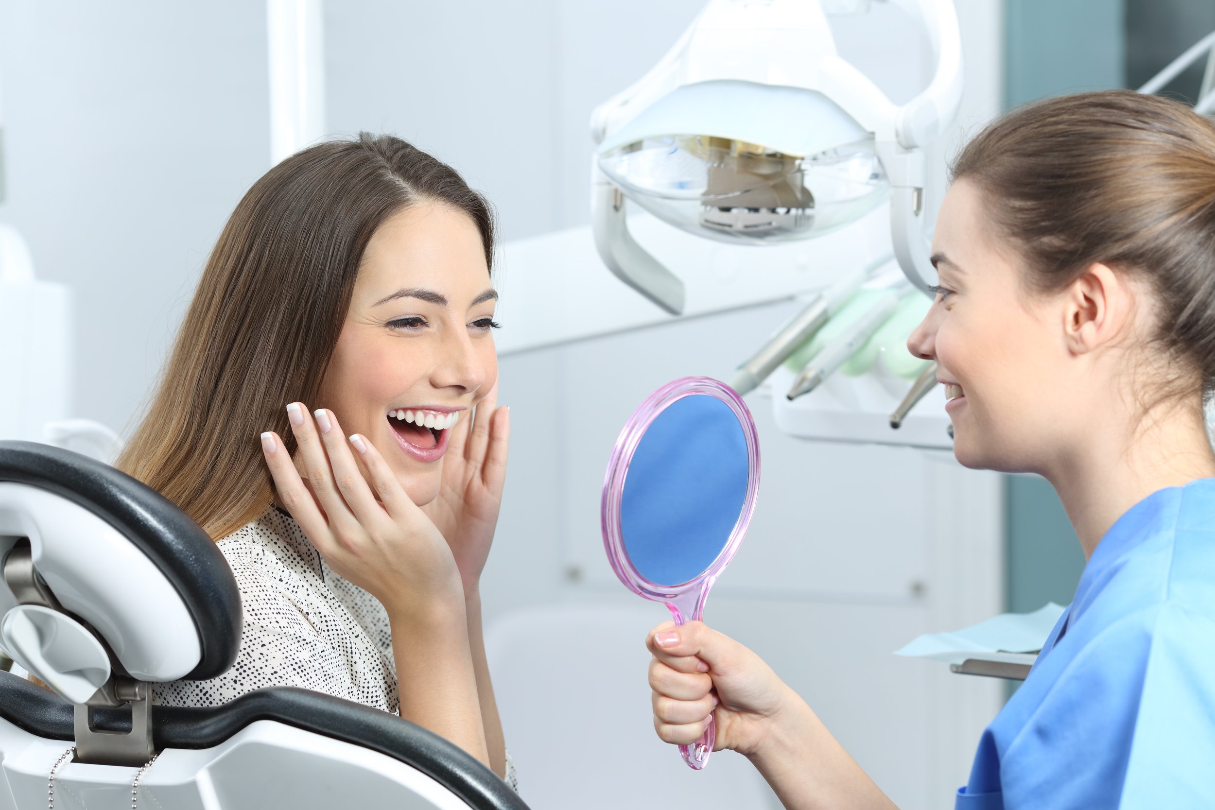 A woman in a dental chair smiling and touching her face, while another woman in dental scrubs holding a pink mirror faces her, in a dental clinic.