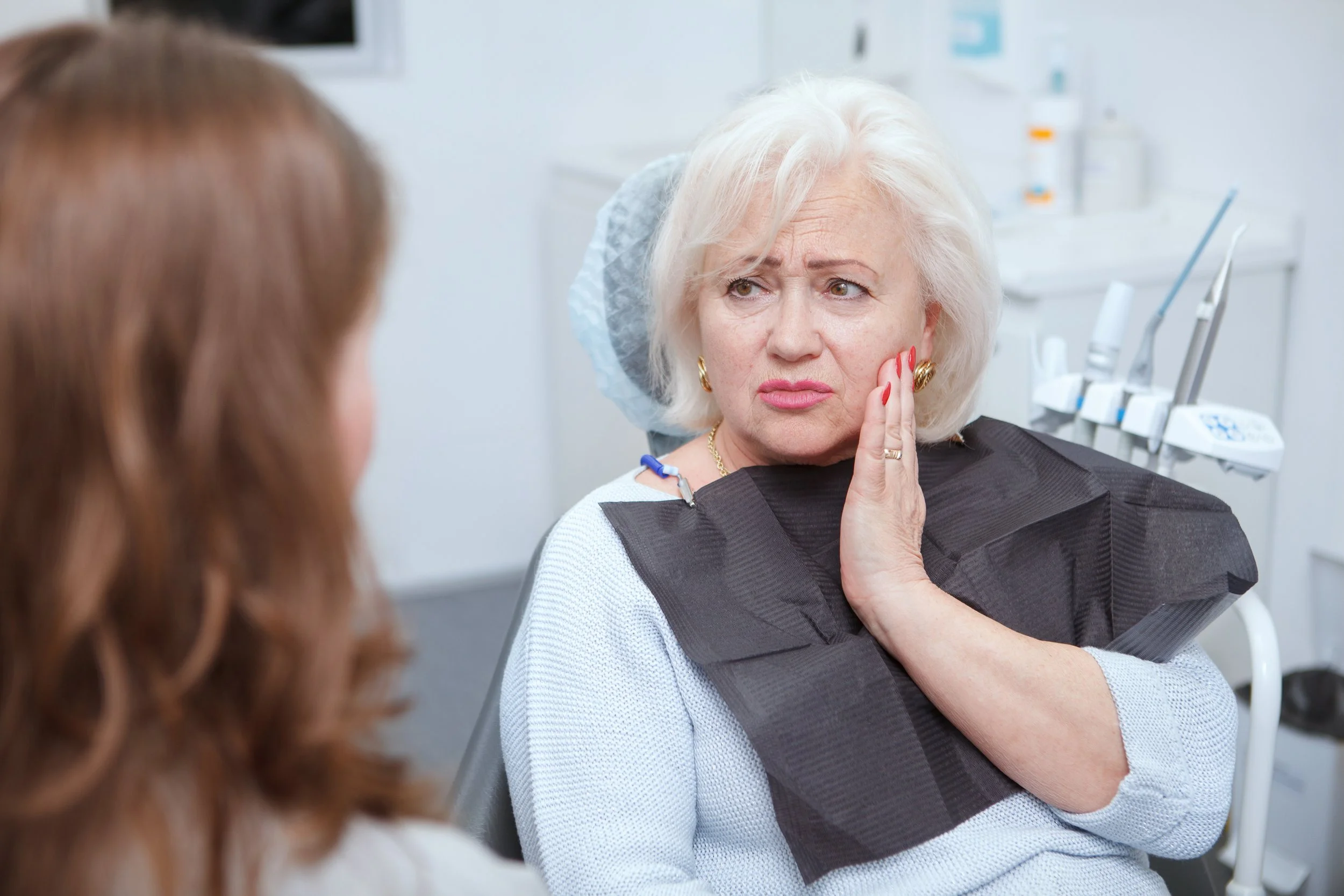 An elderly woman with white hair sitting in a dentist's chair, touching her cheek with a worried expression. A dental assistant or another person is partially visible in the foreground.