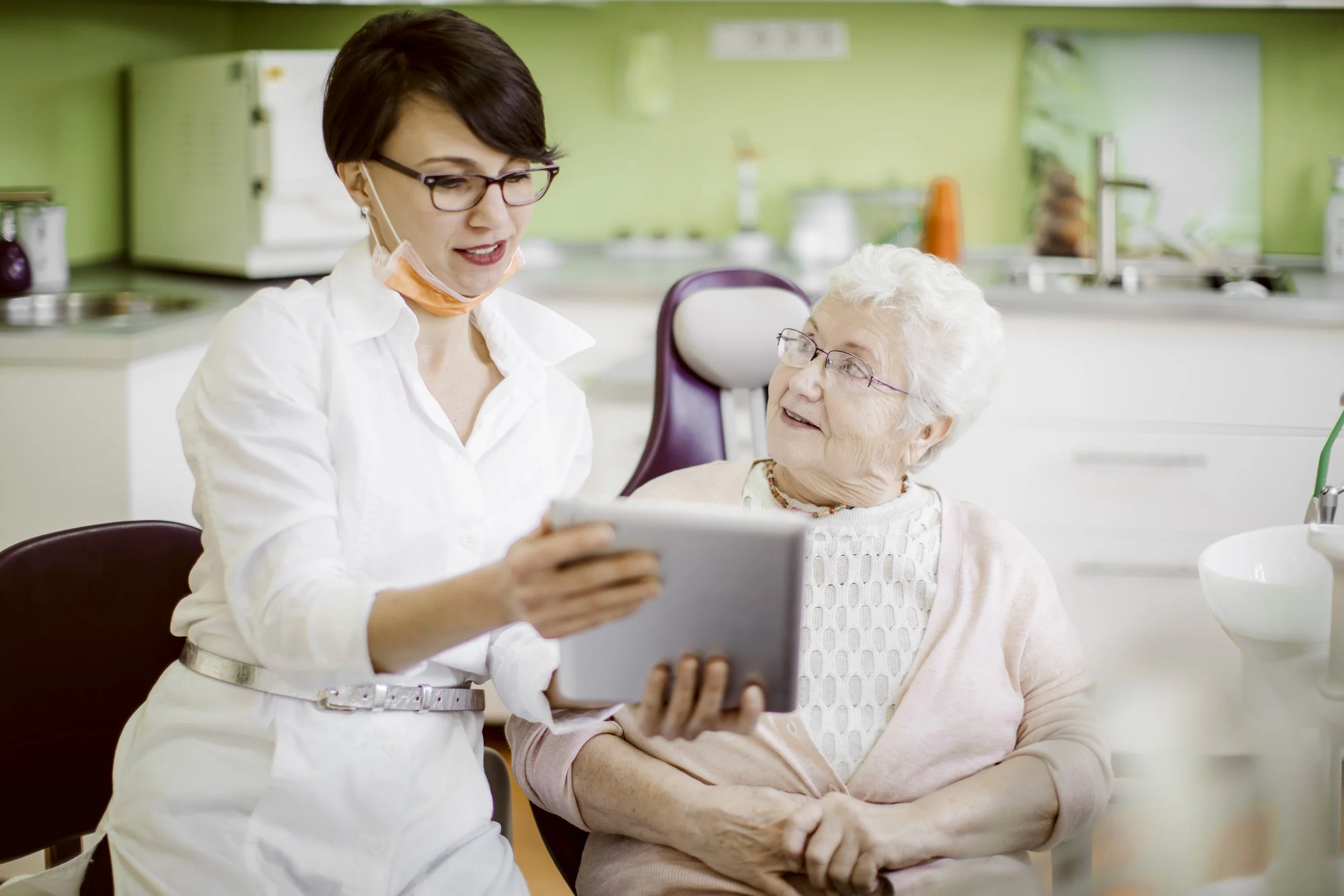 A female healthcare worker shows a tablet to an elderly woman in a bright, clean medical or care facility, with kitchen appliances visible in the background.