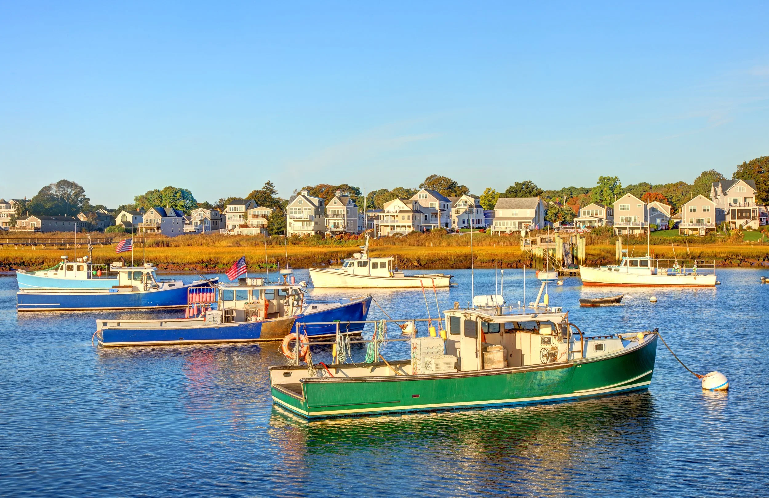 Boats floating on a calm water body with a view of colorful houses on the shoreline, under a clear blue sky.