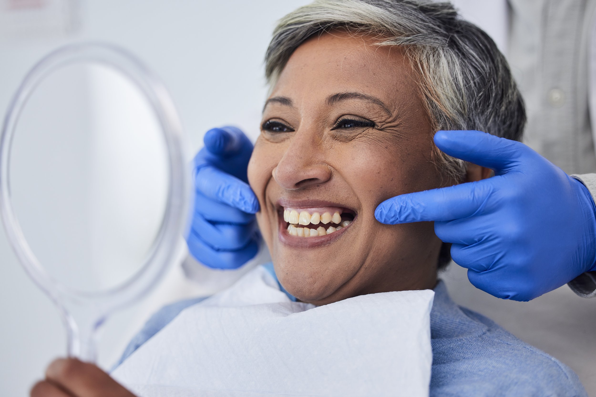 An older woman smiling at her reflection in the mirror, showing her teeth. A dental professional wearing blue gloves is gently touching her face near her cheeks.