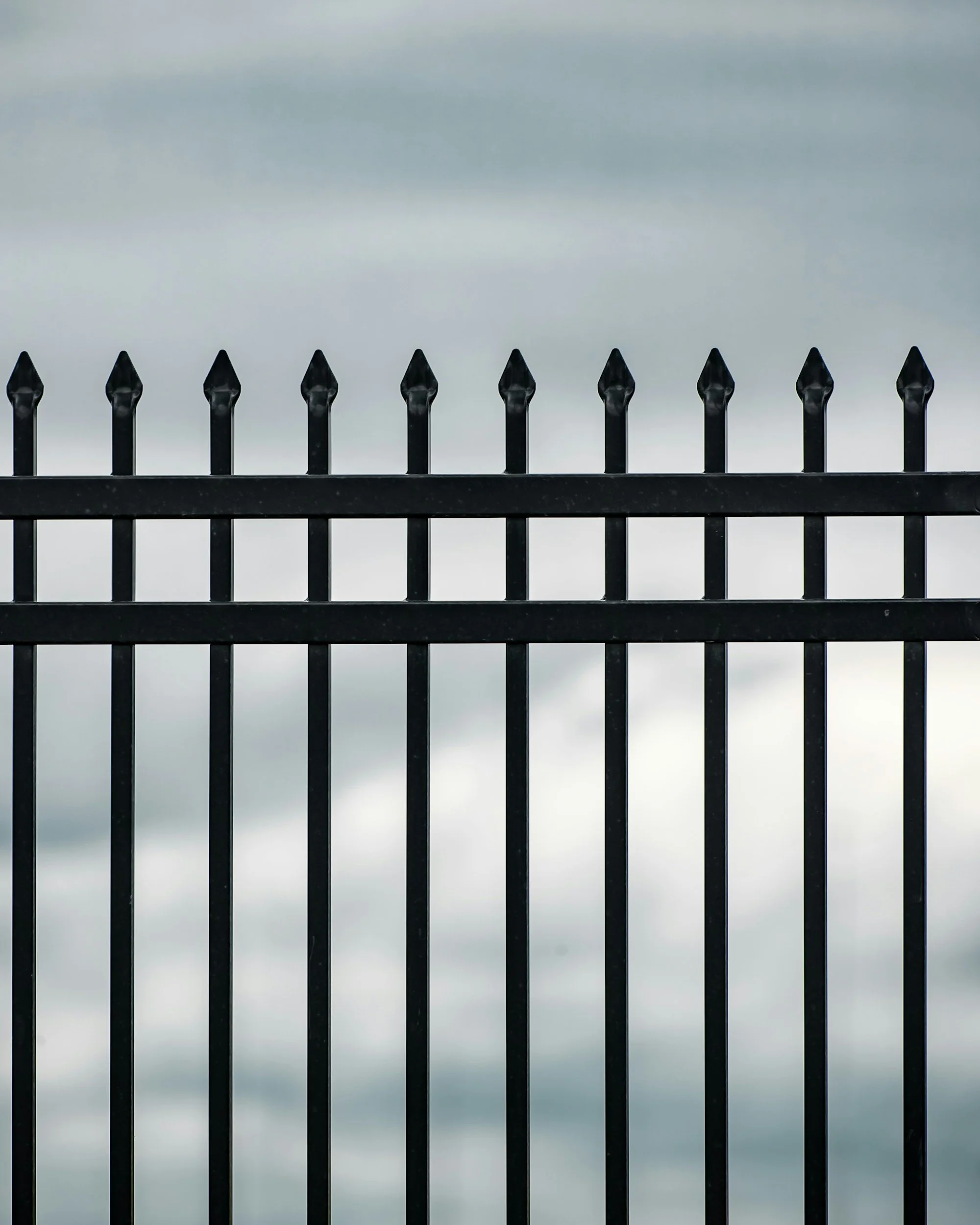 Black metal fence with pointed tips against a cloudy sky.
