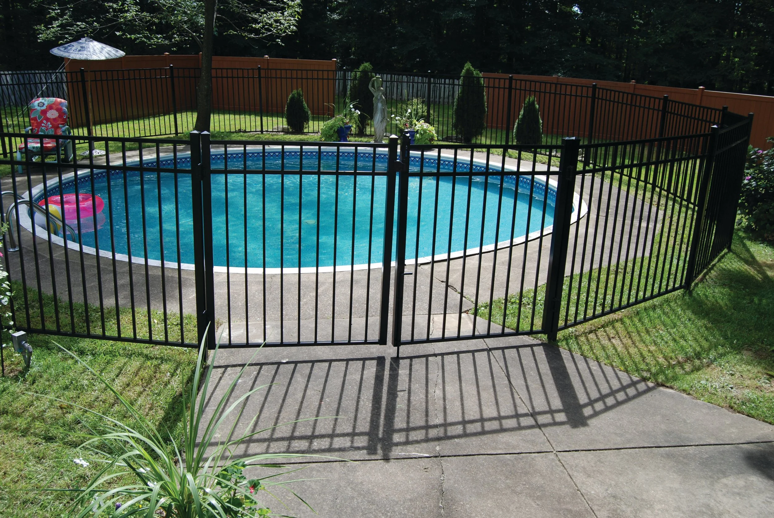 Backyard with a fenced-in swimming pool, surrounded by green grass, potted plants, and decorative statues, with a brown privacy fence and trees in the background.