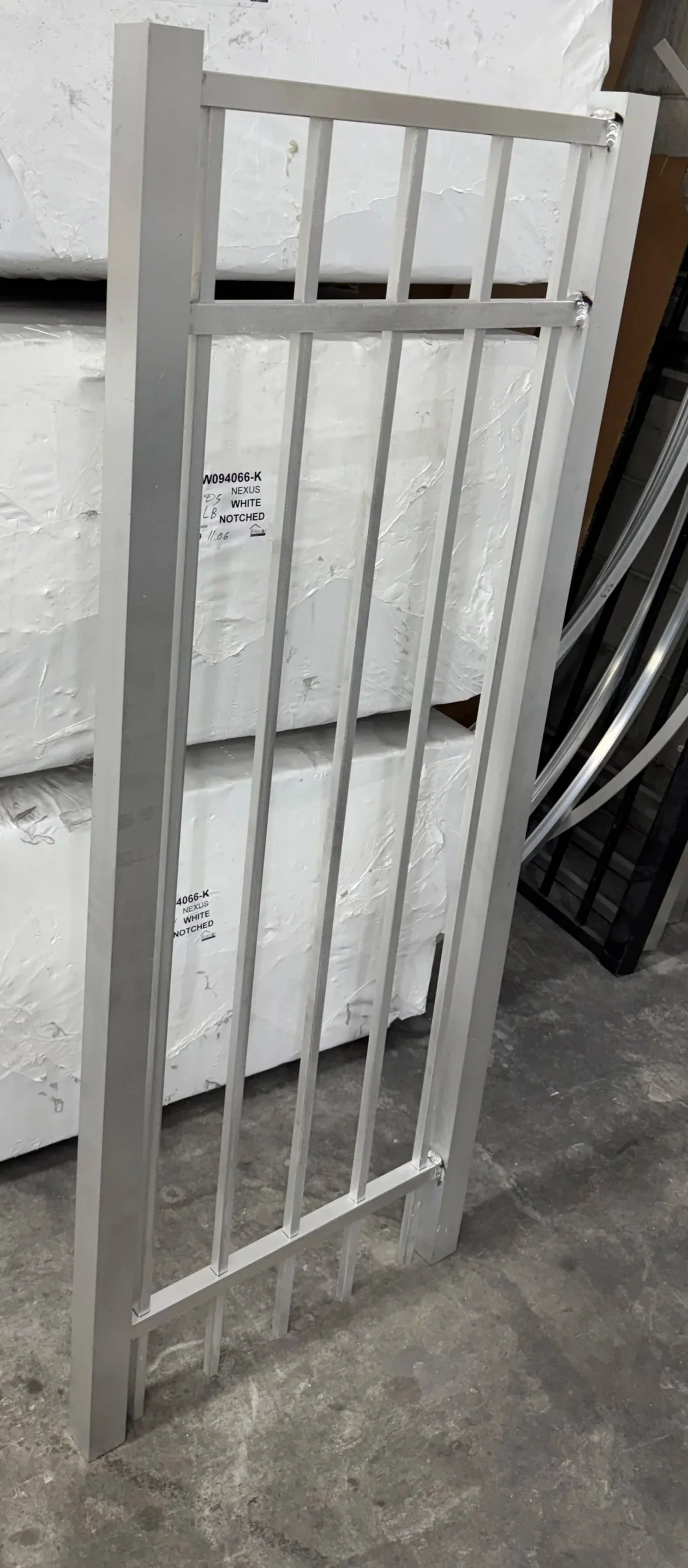 A silver metal child safety gate with vertical bars leaning against a stack of boxed mattresses in a warehouse.