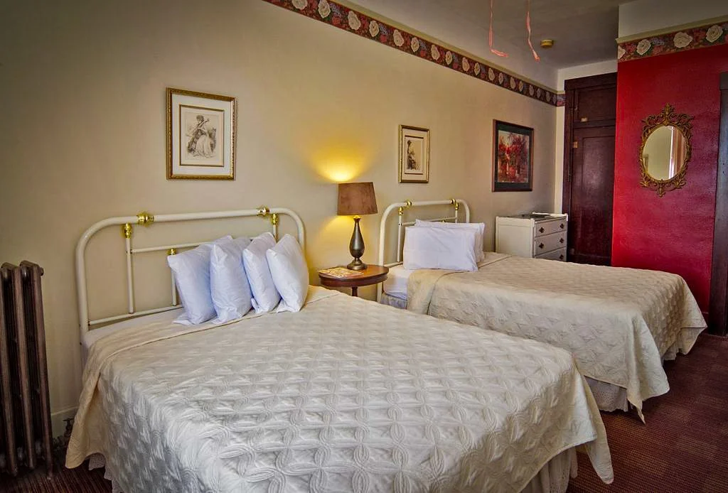 Two beds with white bedding and multiple pillows in a hotel room, with a bedside lamp, framed artwork, and a mirror on the wall.