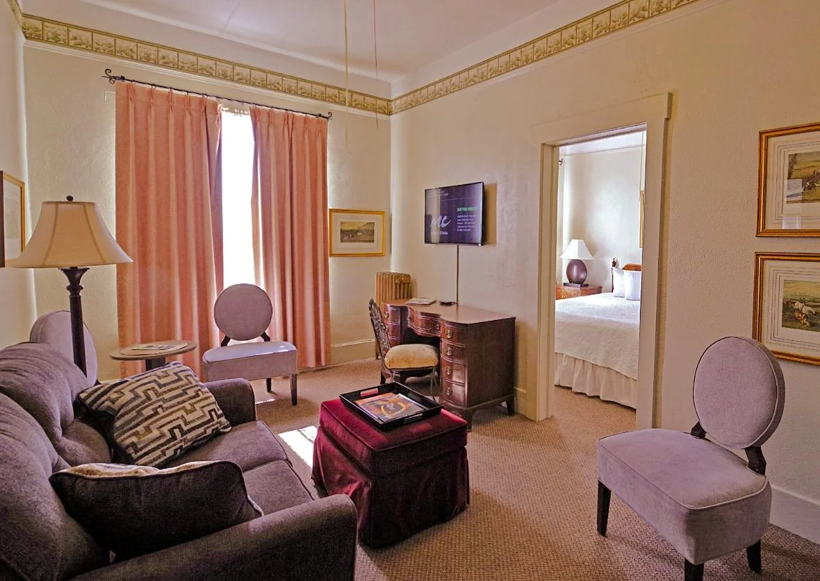 Hotel room living area with a sofa, armchairs, a desk with a television, and a nearby bedroom with a bed and nightstand.