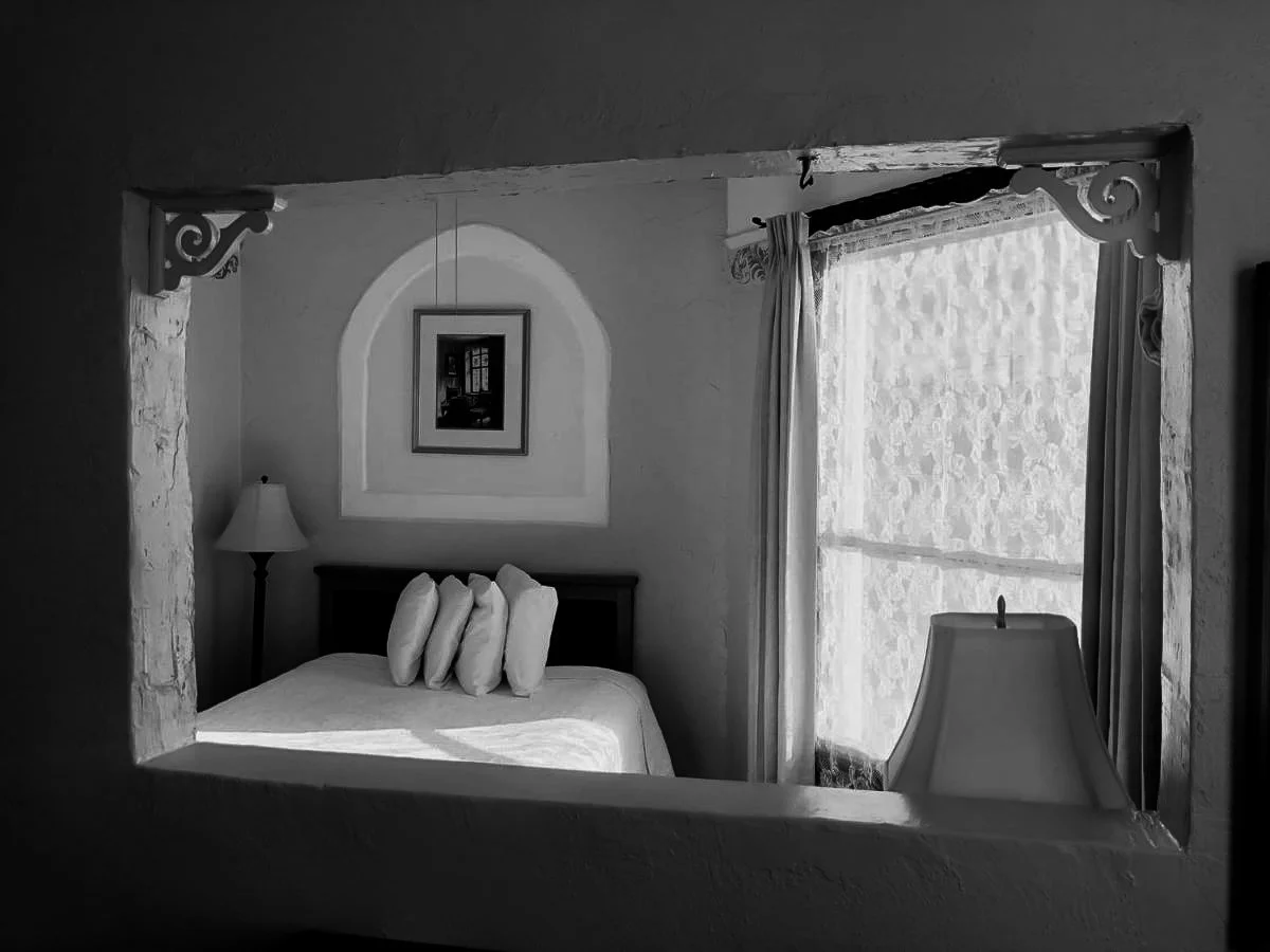 Black and white photo of a bedroom viewed through a window, showing a bed with pillows, a lamp, framed picture on the wall, and sunlight coming through sheer curtains.