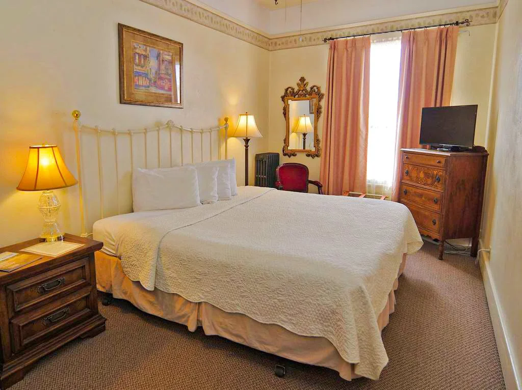 A hotel room with a large bed, nightstands with lamps, a dresser with a TV, a mirror, a framed picture on the wall, pink curtains, and a chair.
