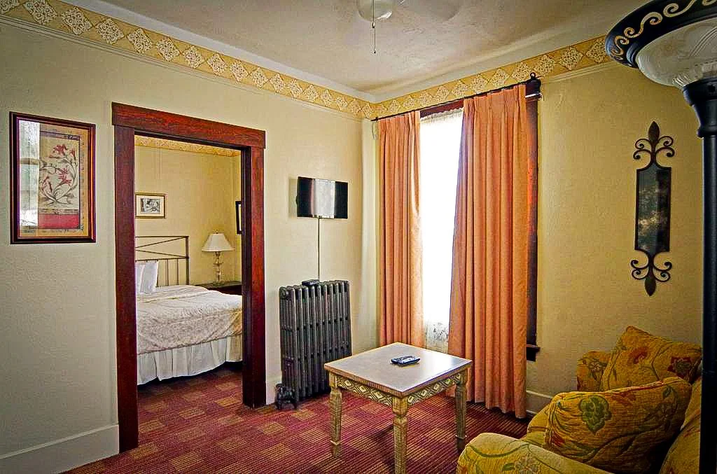 A cozy hotel room with a yellow-patterned armchair, a small ornate coffee table, a window with peach curtains, and a doorway leading to a bedroom with a bed and nightstand.