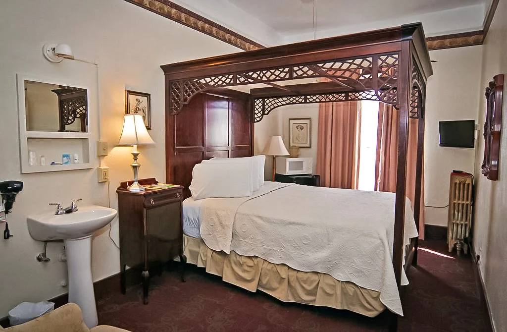 Hotel room with a four-poster bed, nightstand, lamps, a small TV, and a sink.
