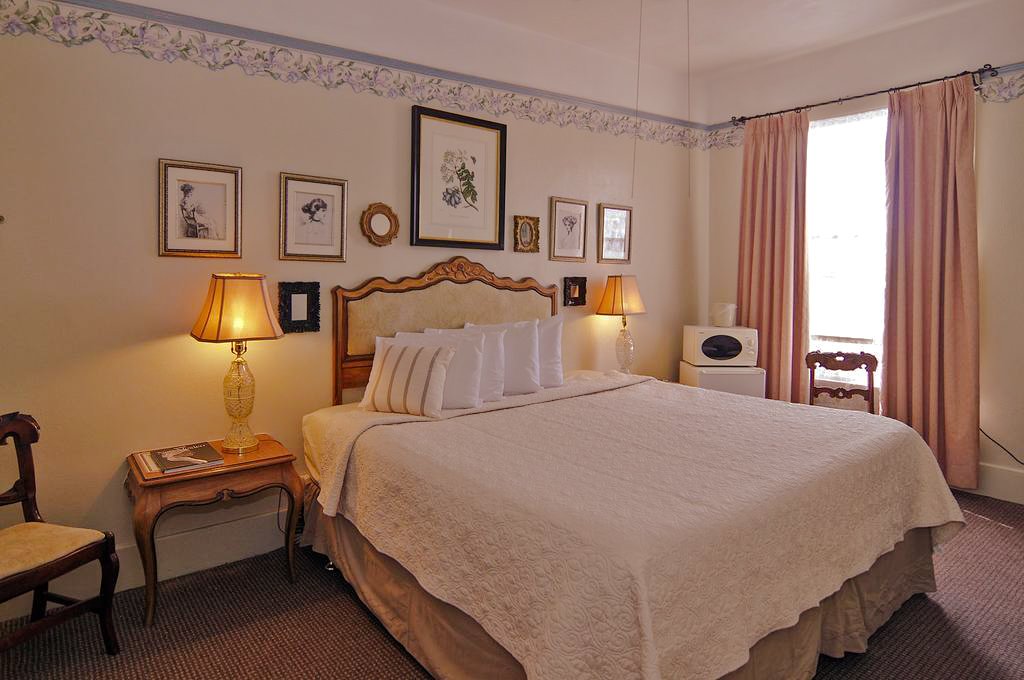 A cozy bedroom with a large bed, decorative frame above headboard, and bedside lamps with warm lighting. The wall features framed art, and there are curtains covering a window, with a small white cabinet and a chair nearby.