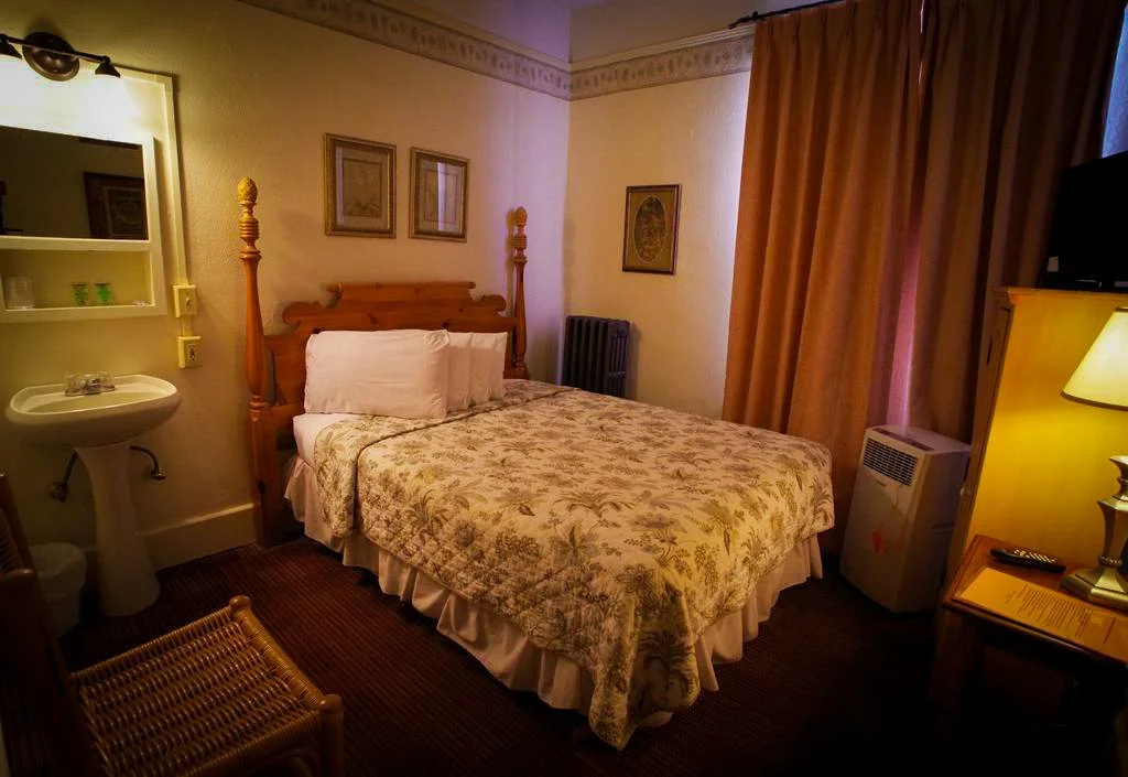 Small hotel room with a bed, wooden headboard, pillows, floral bedspread, a small sink with mirrored cabinet, framed pictures on the wall, window with curtains, and various furniture including a nightstand, lamp, TV, and heater.