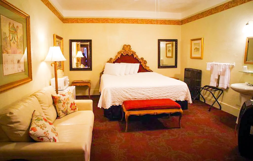 A cozy hotel room with a large bed, decorative headboard, sofa with floral pillows, bedside lamps, a mirror, framed artwork, a small side table, a vintage bench, a radiator, a towel rack, a sink, and a small mirror.