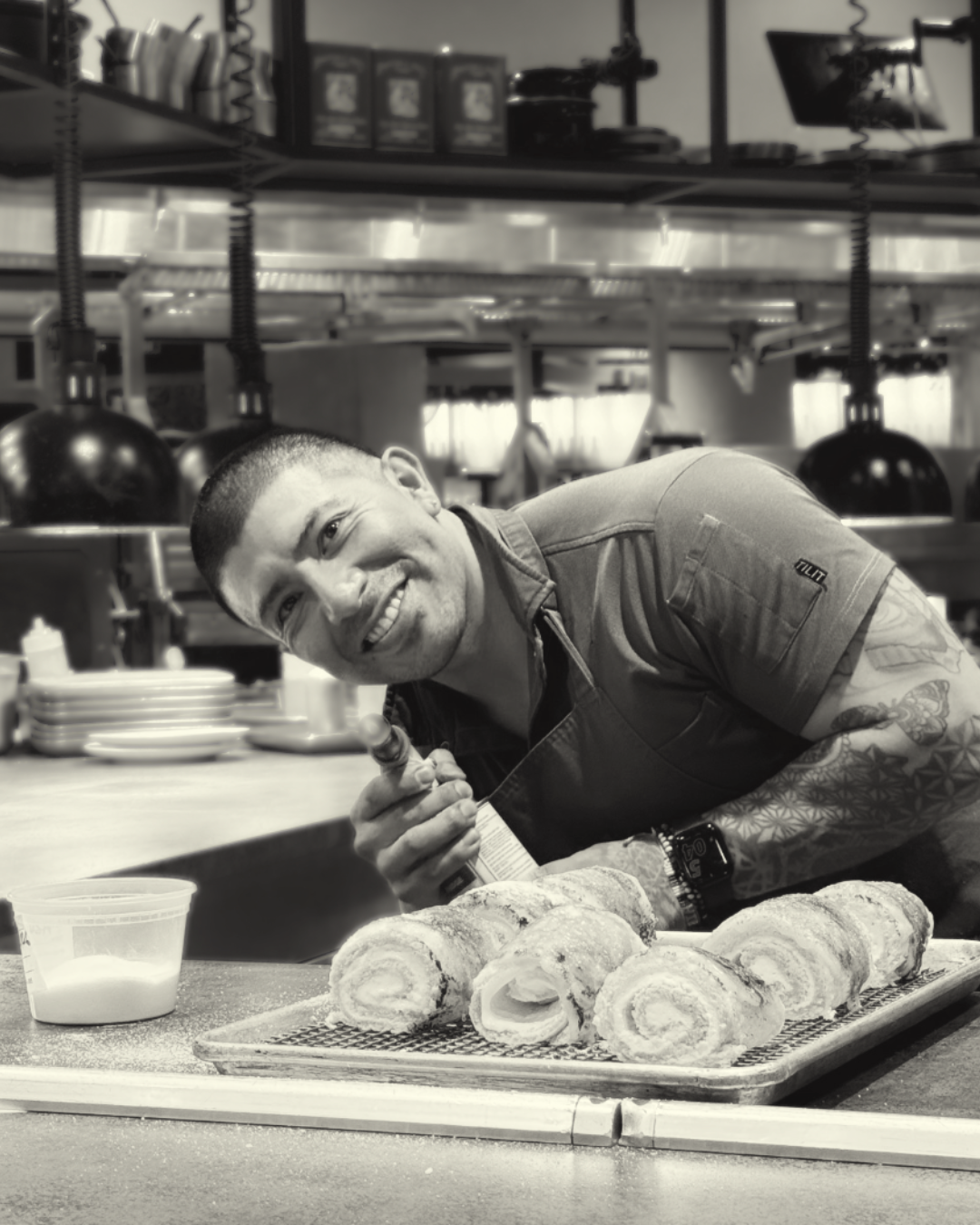 A smiling man with tattoos on his right arm, holding a spoon, leaning over a tray of rolled pastries in a kitchen or restaurant setting.
