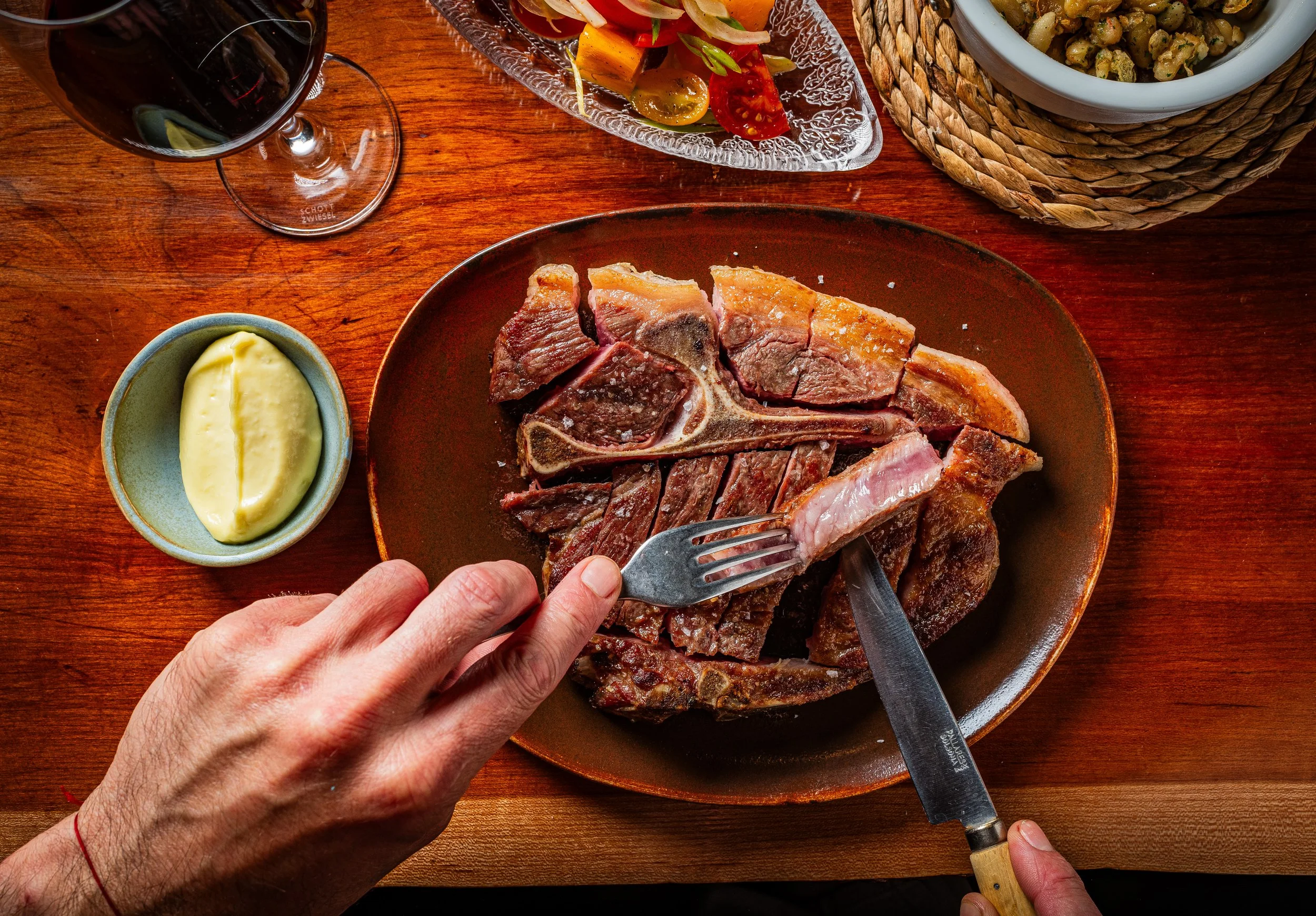 A person cutting a cooked beef steak on a brown plate, with a fork in hand, on a wooden table. Nearby are a small bowl of butter, a glass of red wine, a bowl of salad with cherry tomatoes, and a covered dish with a vegetable mixture.