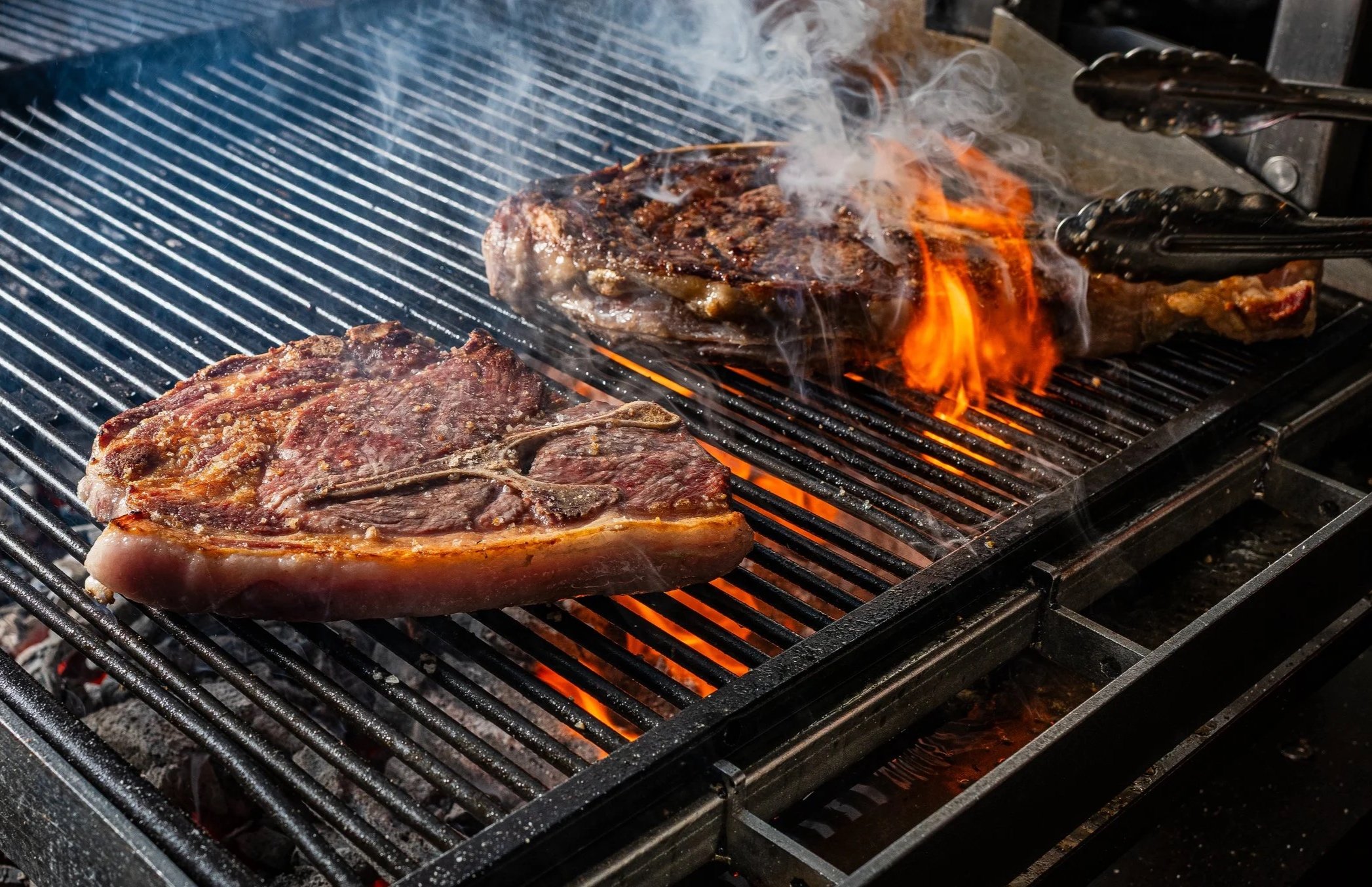 Two large beef steaks grilling on a barbecue with flames and smoke.