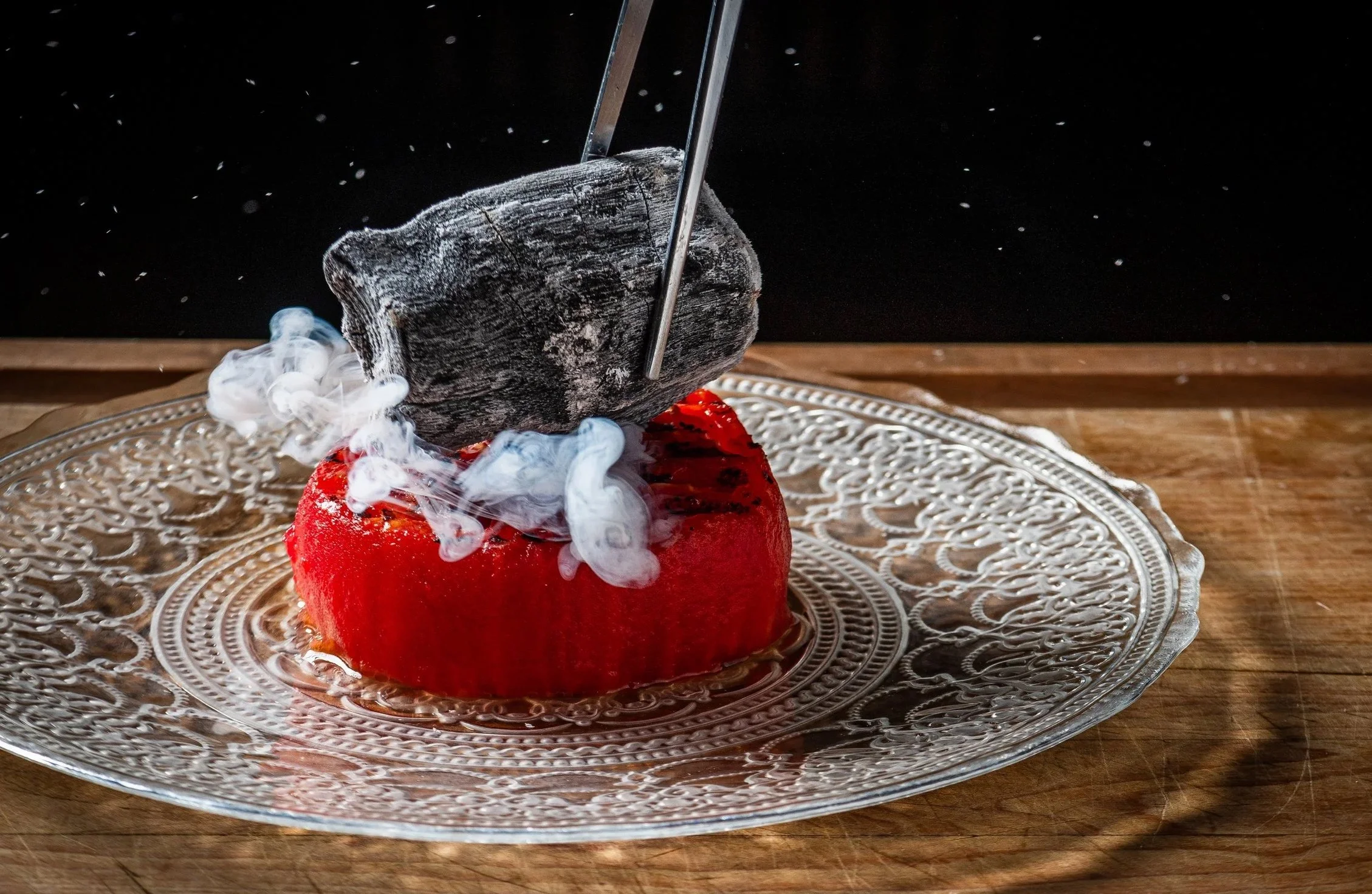 A red dessert topped with a smoky, dark object being smoked with smoldering wood or herbs, on a decorative glass plate on a wooden surface against a black background.