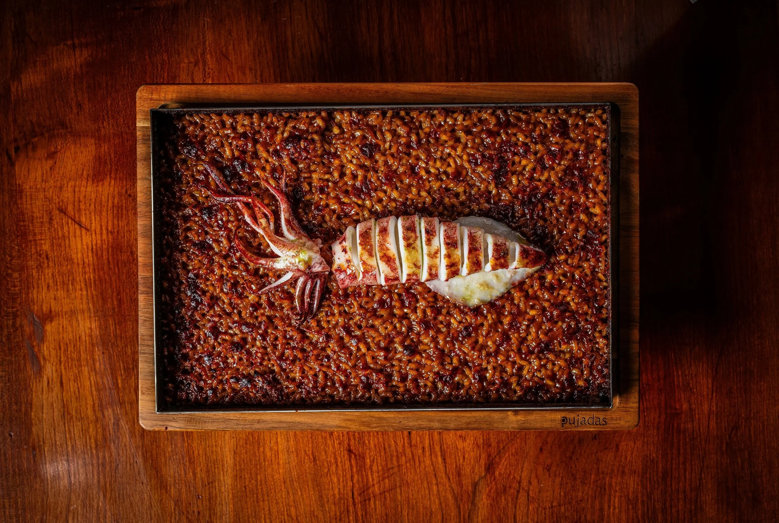 A rectangular wooden tray filled with cooked, seasoned rice and sliced grilled squid, placed on a dark wooden surface.