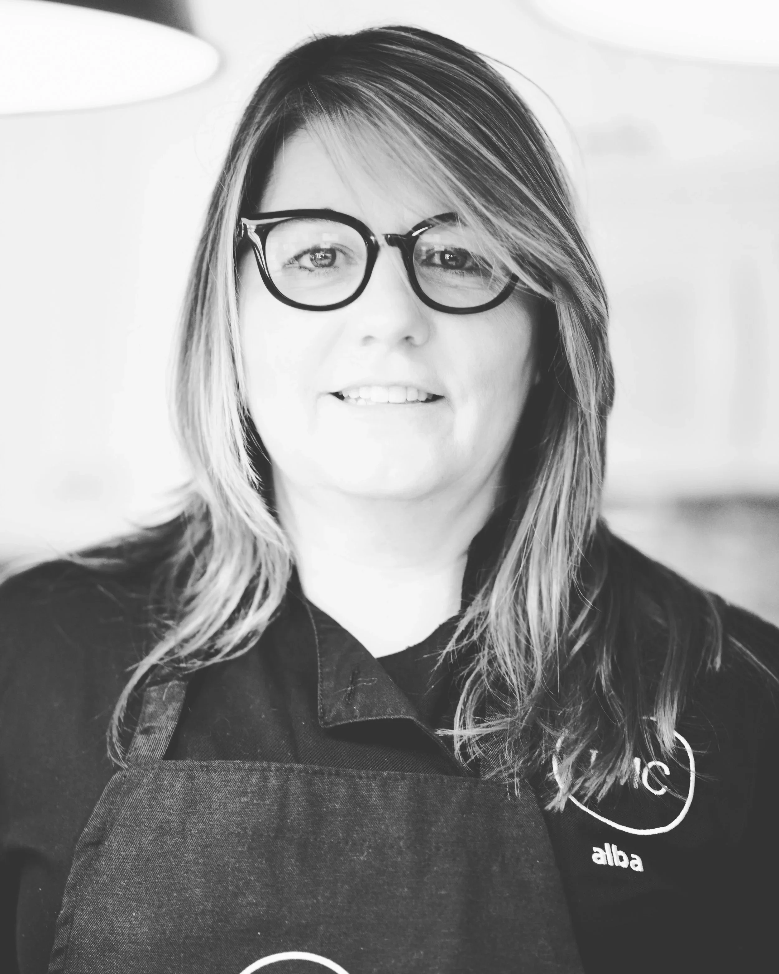 A woman with shoulder-length hair wearing glasses and a dark shirt. She is smiling slightly and looking directly at the camera.