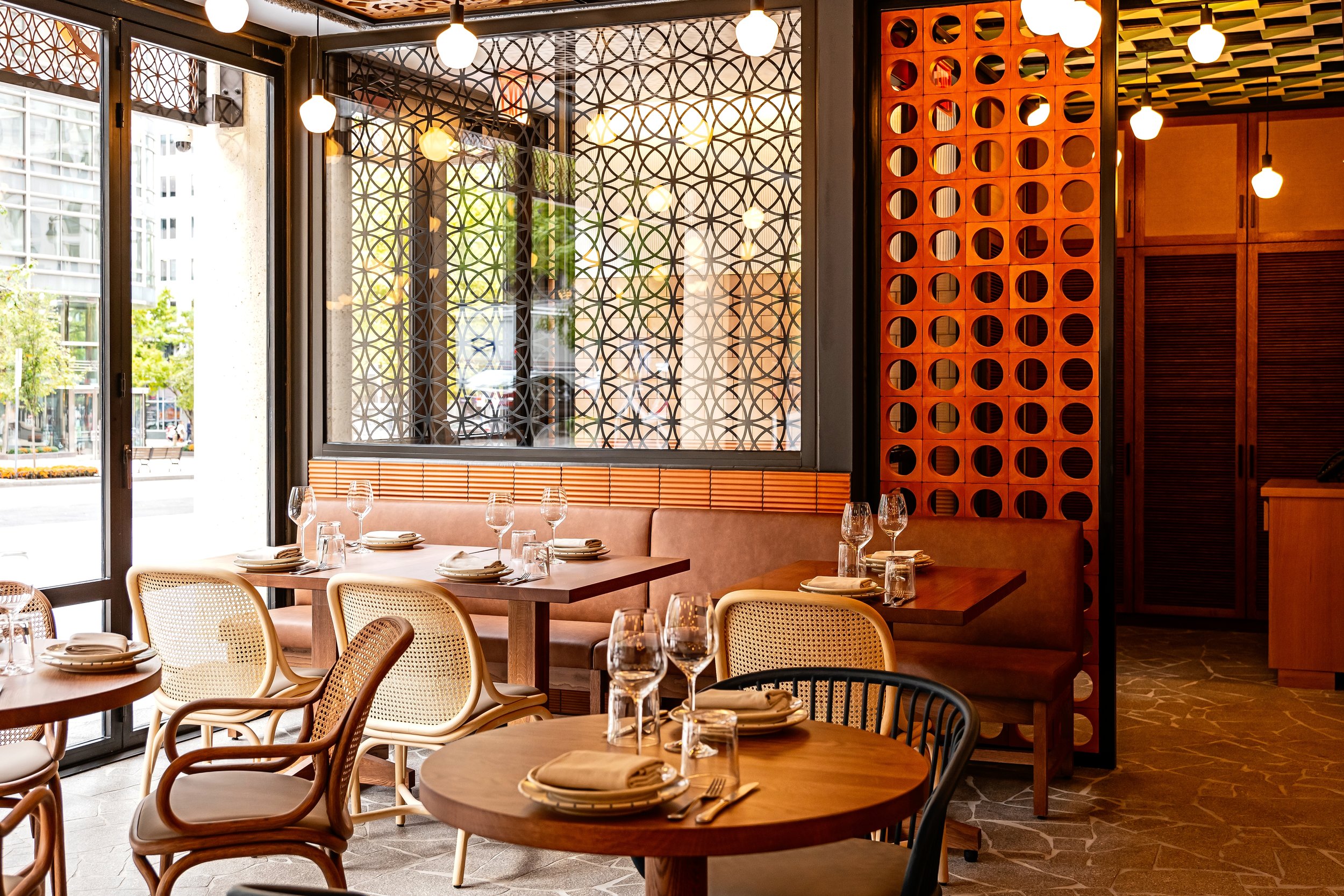 Interior of a restaurant with round wooden tables set with plates, napkins, forks, and wine glasses. Light bulbs hang from the ceiling, and there are large windows with decorative metal grille panels. The decor features warm wood tones and geometric 