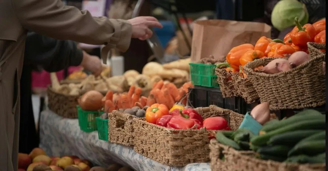 A market stall with various fresh vegetables in baskets, including bell peppers, zucchinis, carrots, onions, and garlic, with people shopping.