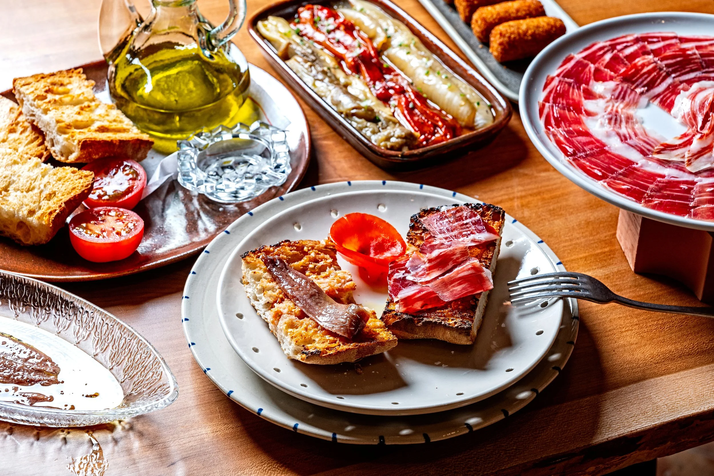 A variety of Spanish tapas dishes on a table, including slices of bread with toppings, cherry tomatoes, olive oil, roasted vegetables, and cured meats.