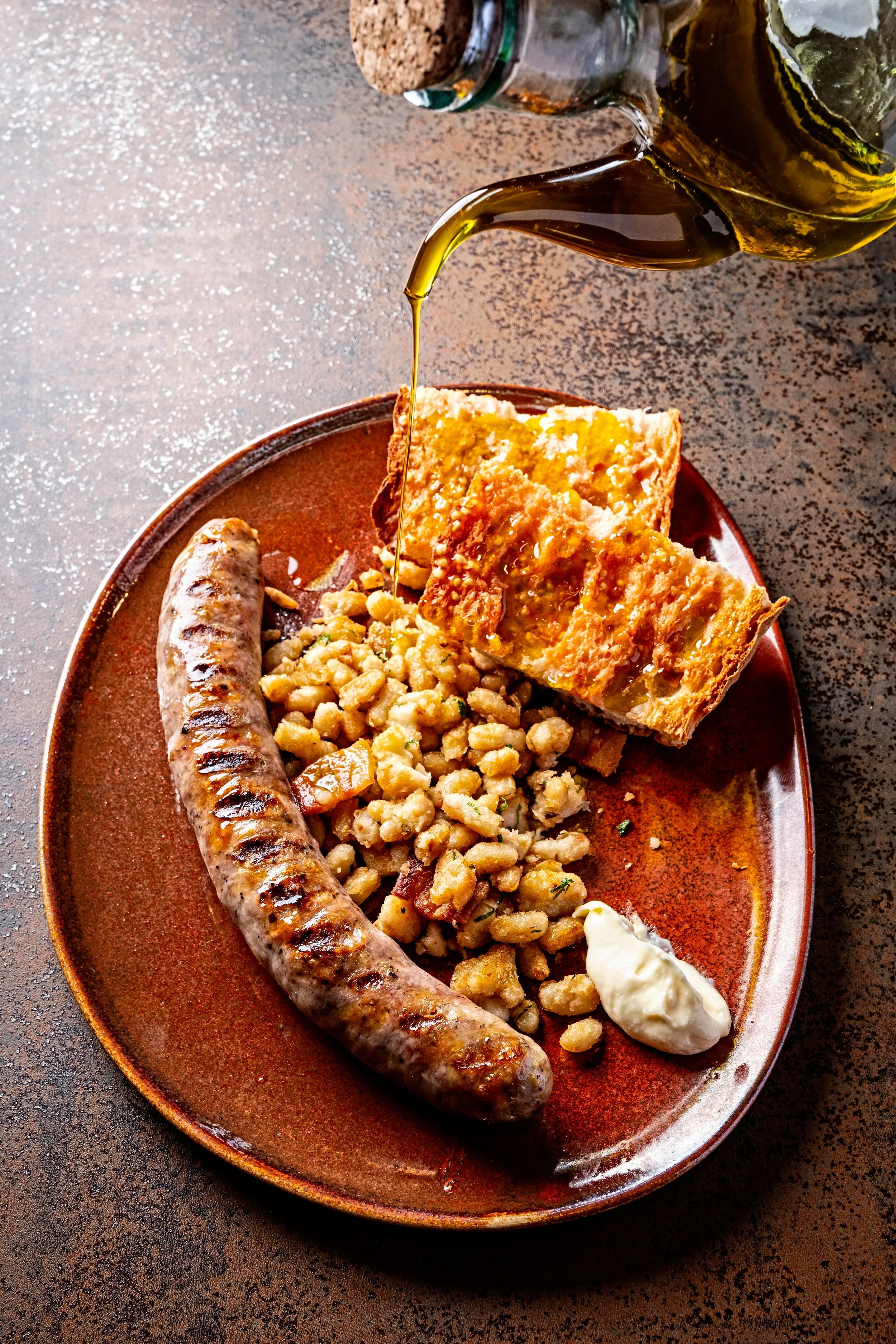 Sausage with grill marks on a brown plate, served with a large slice of cornbread with butter, a scoop of white sauce, and some potato salad, with a bottle of syrup on a textured surface.