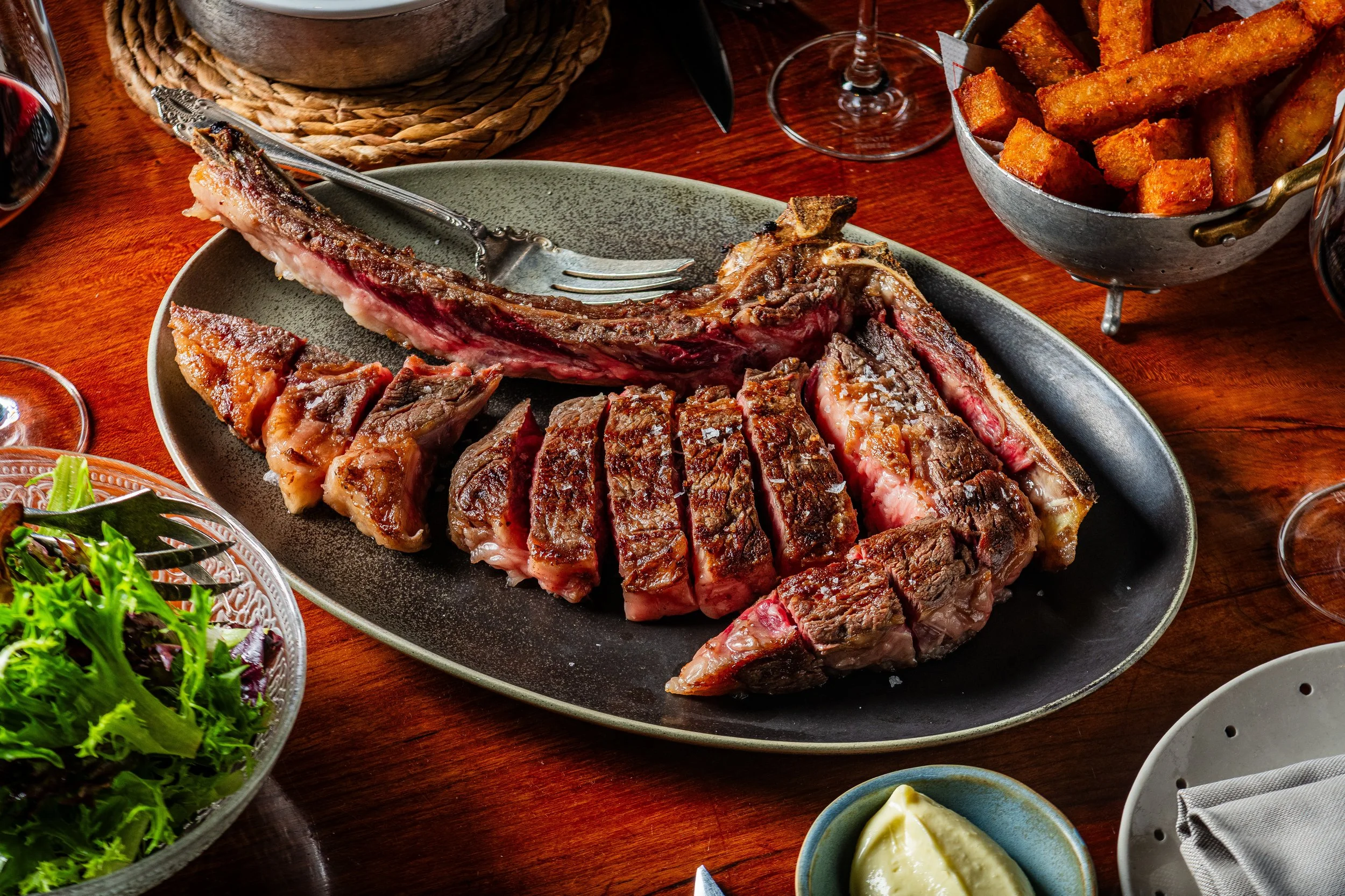 A plate of cooked steak, sliced and with a bone, on a dining table with various side dishes and glasses of wine.