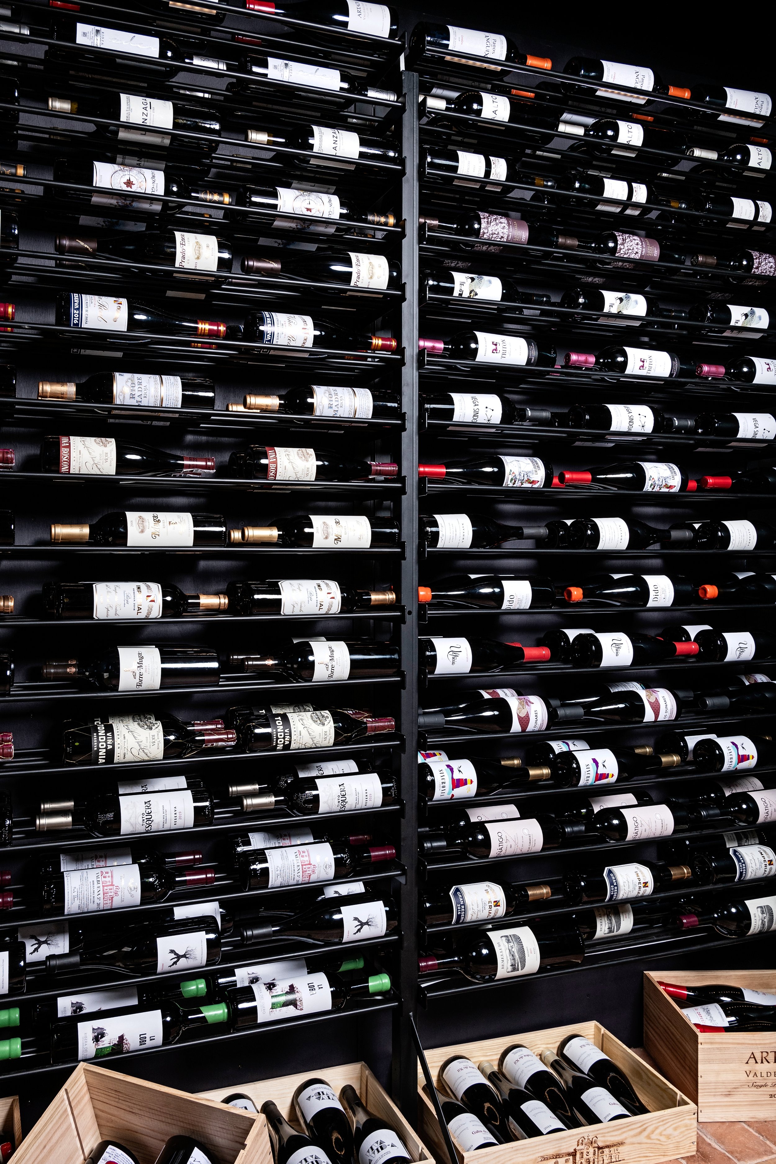 A display of wine bottles organized on black metal racks with wooden crates filled with more bottles at the bottom.